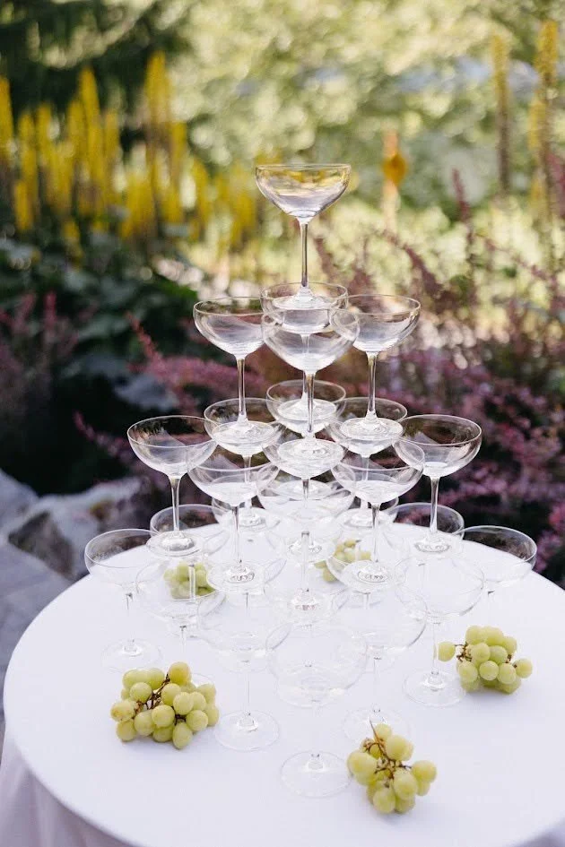 A pyramid arrangement of empty champagne glasses on a white table, with bunches of green grapes nearby, outdoors in a garden setting.