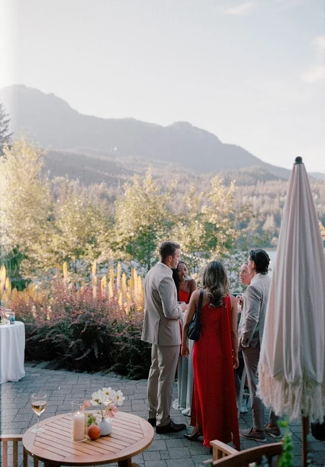 Group of people socializing outdoors in a scenic setting with mountains and trees in the background, some holding drinks.