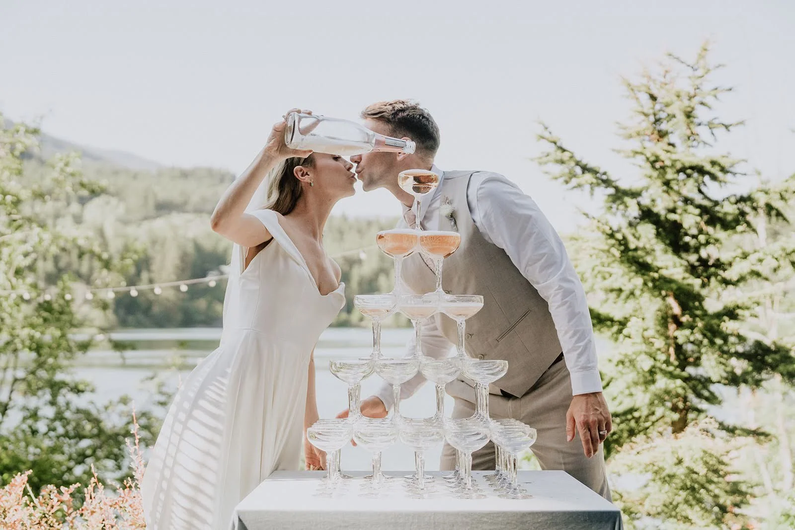 A bride and groom kissing while pouring champagne into a champagne tower at an outdoor wedding reception.