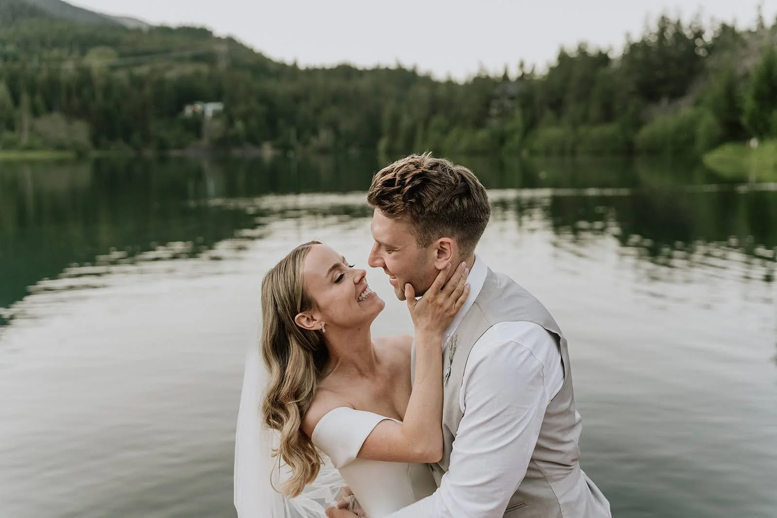 A smiling bride and groom embrace by a lake, with lush green trees and hills in the background.