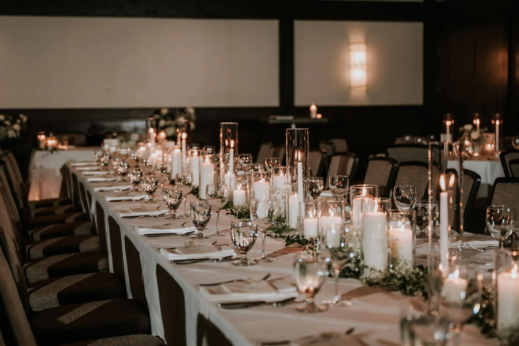 Long dining table decorated with candles, glassware, and greenery in a dimly lit event space.