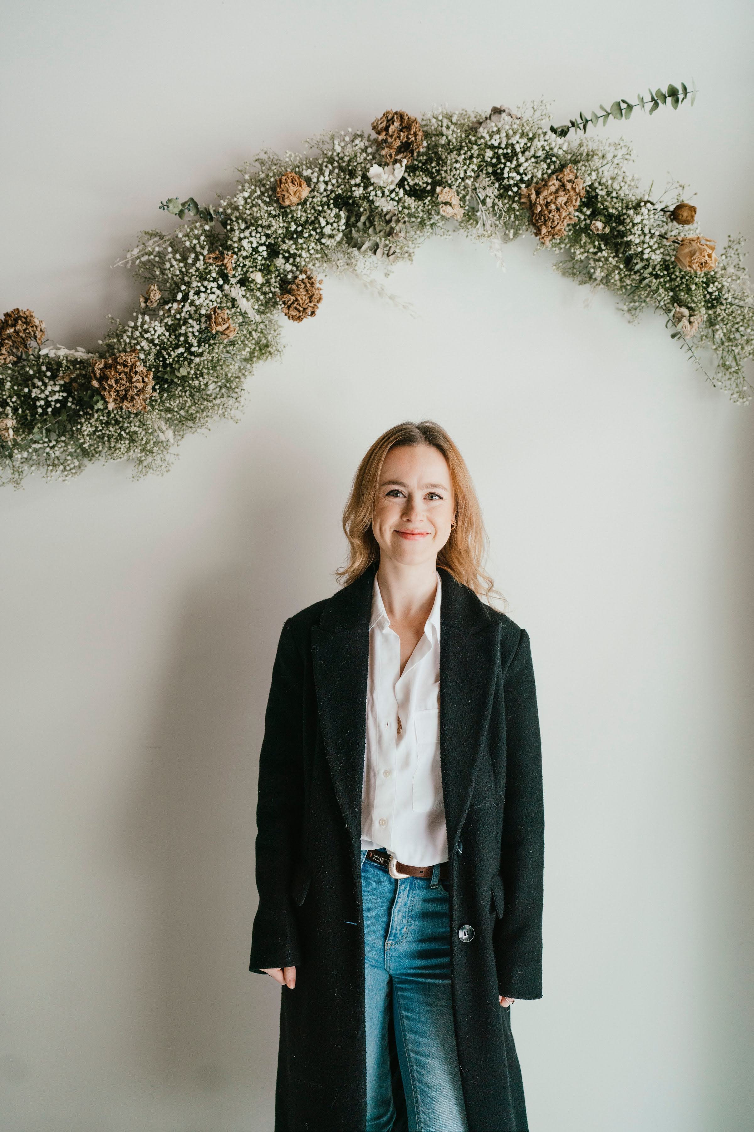 A woman with shoulder-length blonde hair smiling, standing in front of a white wall decorated with a floral arch of white and brown flowers and greenery.