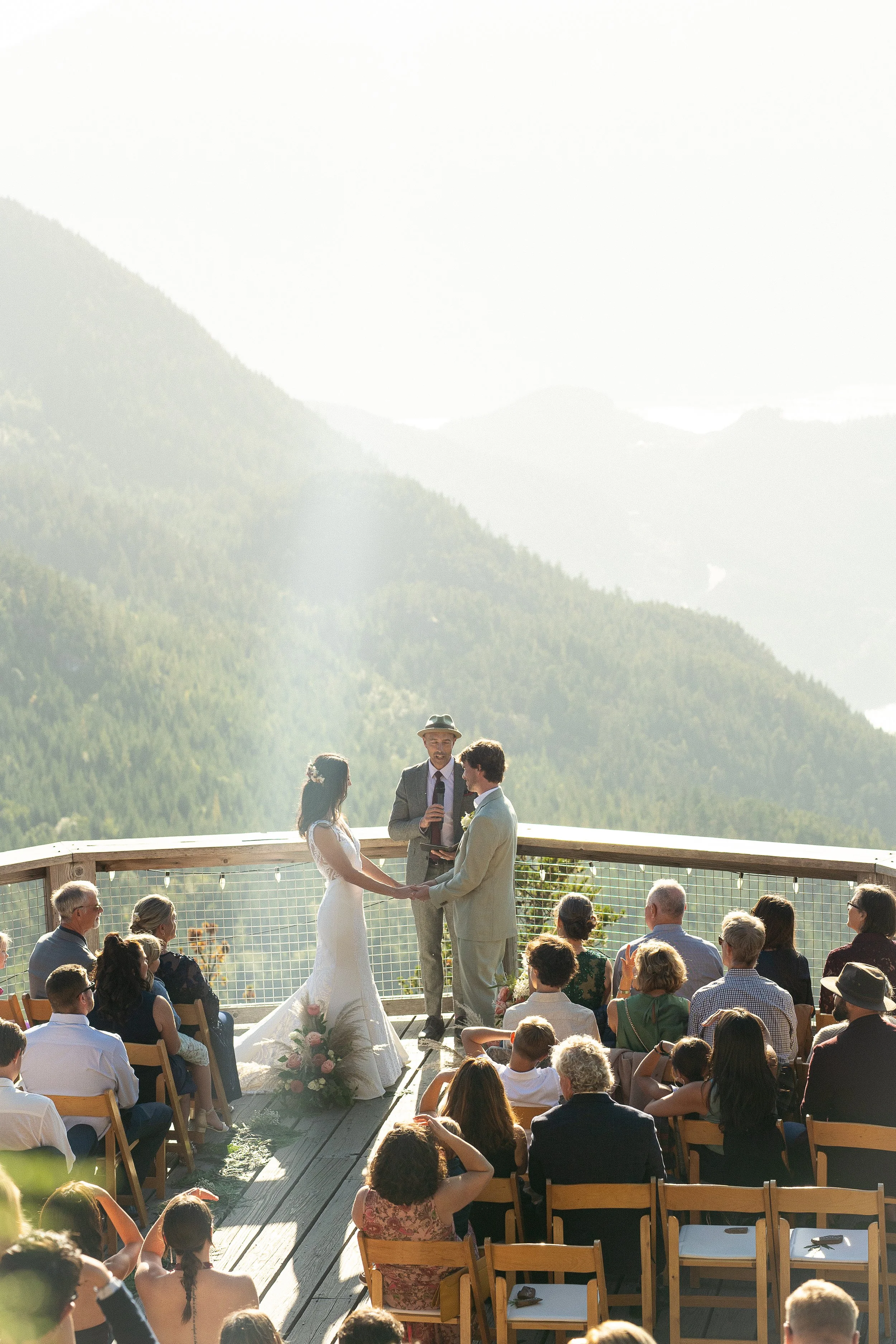 A wedding ceremony outdoors on a platform overlooking mountains, with a bride and groom holding hands, officiant with a microphone, and seated guests watching.