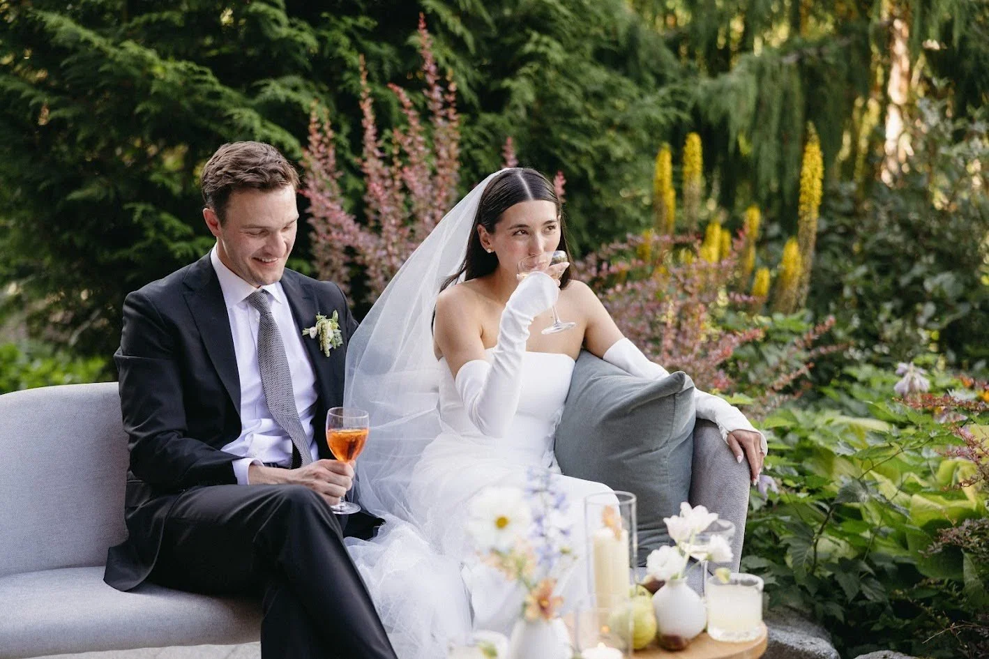 A bride and groom sitting on a outdoor sofa, drinking cocktails, surrounded by lush greenery and flowers, during their wedding celebration.