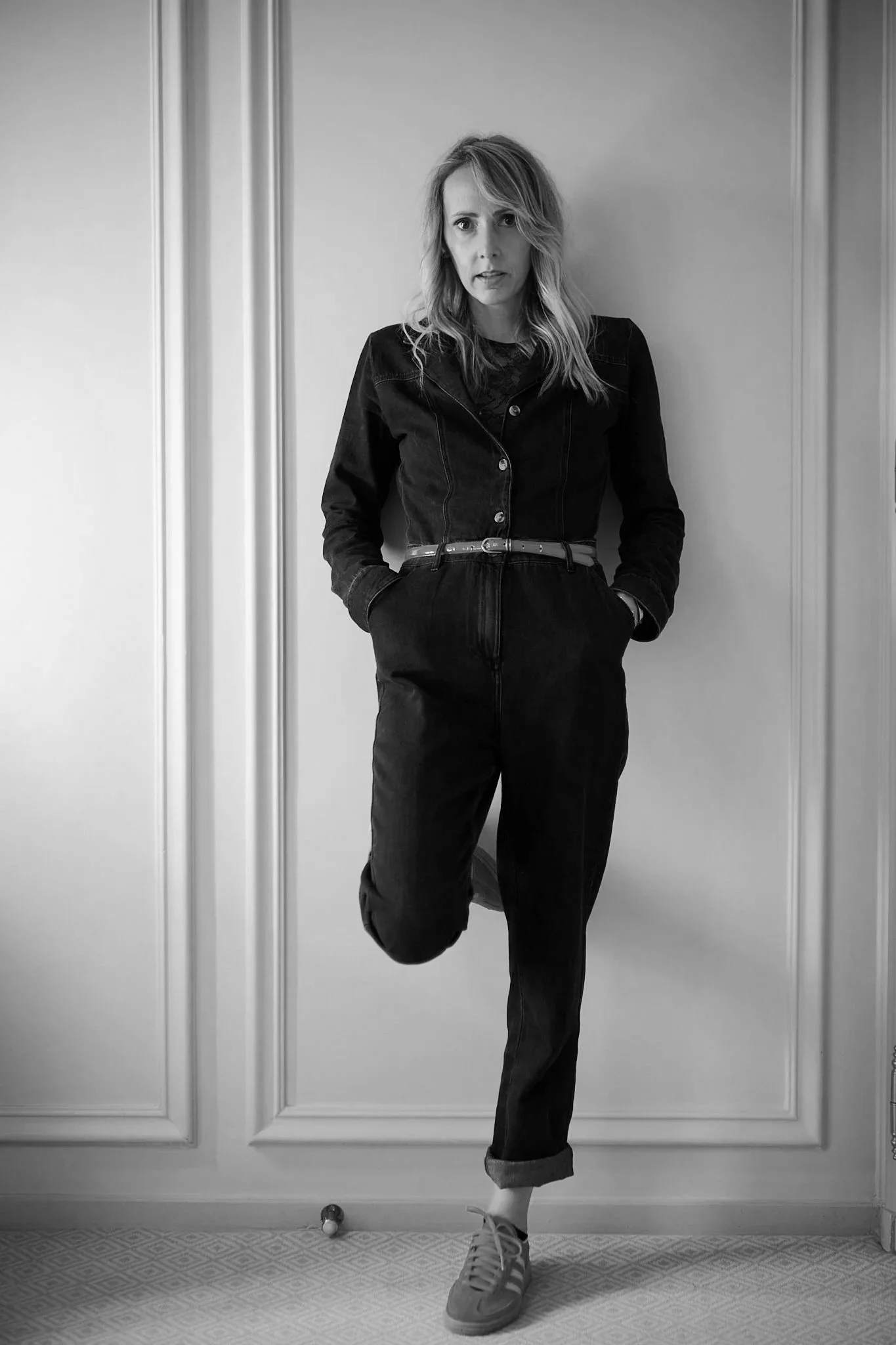 A woman standing indoors against a plain wall with her hands in her pockets, wearing a dark denim jacket, rolled-up jeans, and sneakers, looking at the camera with a serious expression.