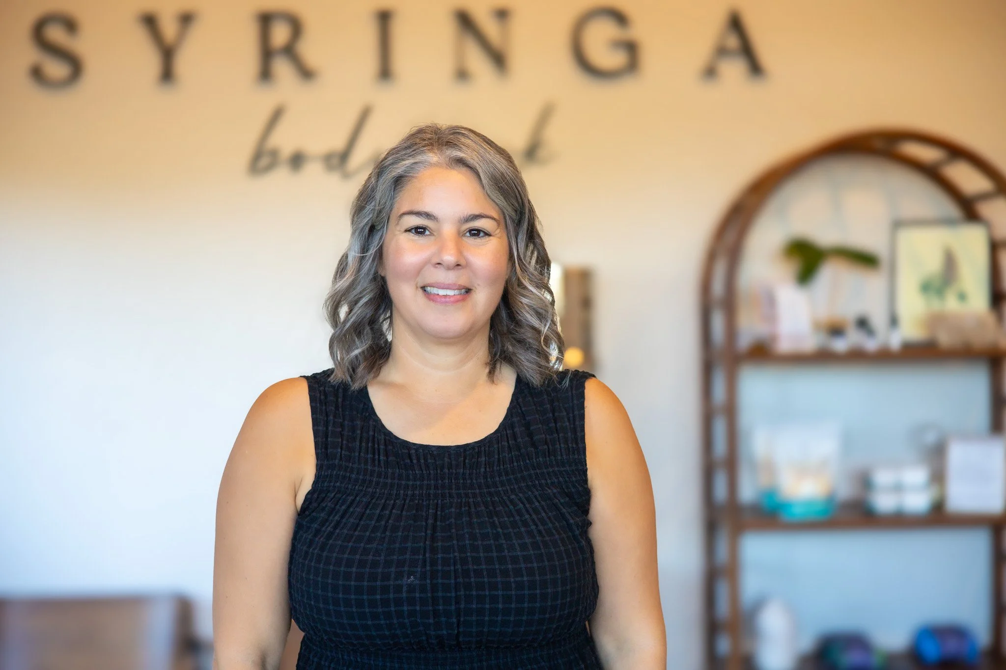 A woman with wavy gray hair smiling at the camera, standing in a wellness or beauty studio with the words 'SYRINGA' and 'body' on the wall behind her.