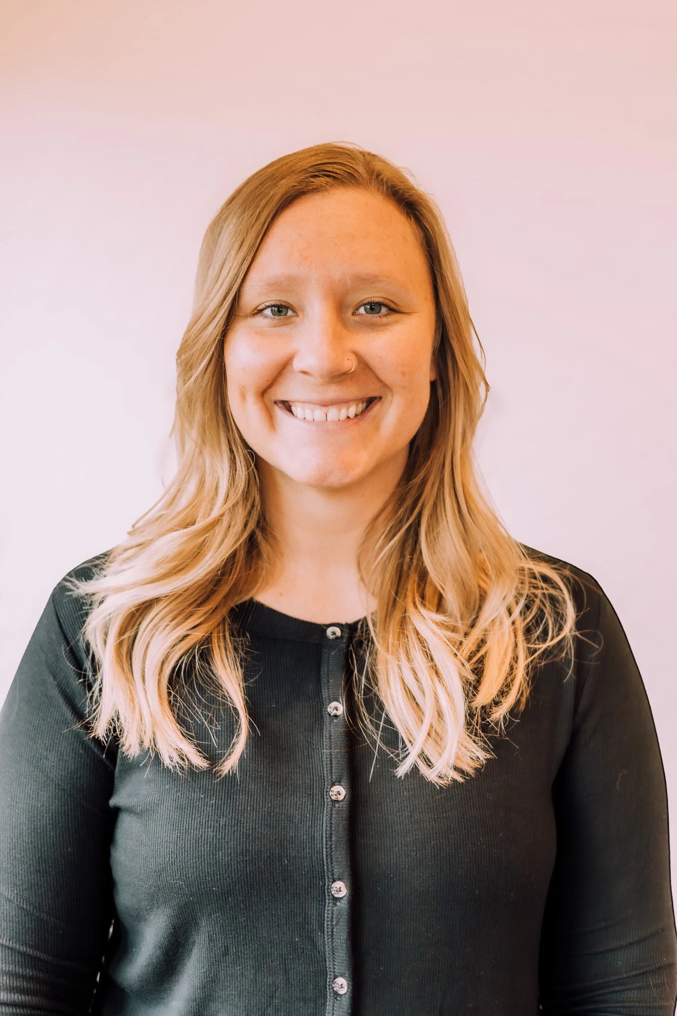 A woman with long, wavy blonde hair, smiling, wearing a black top with buttons, standing against a light pink background.