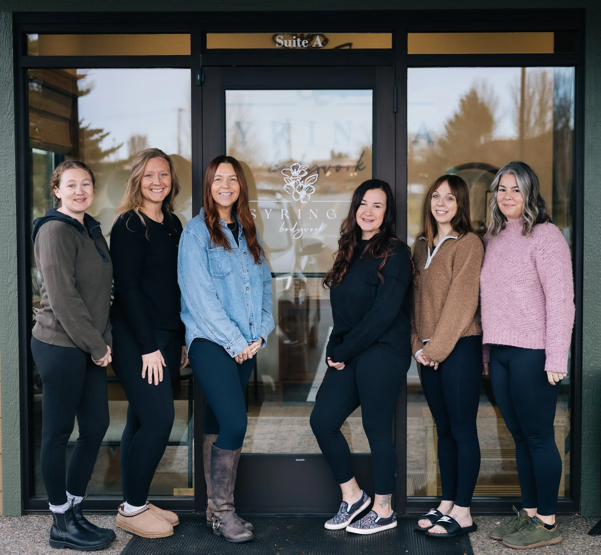Six women standing in front of a glass door with a logo that says 'Syring Bodywork' and a floral design. The women are smiling and dressed in casual clothing.