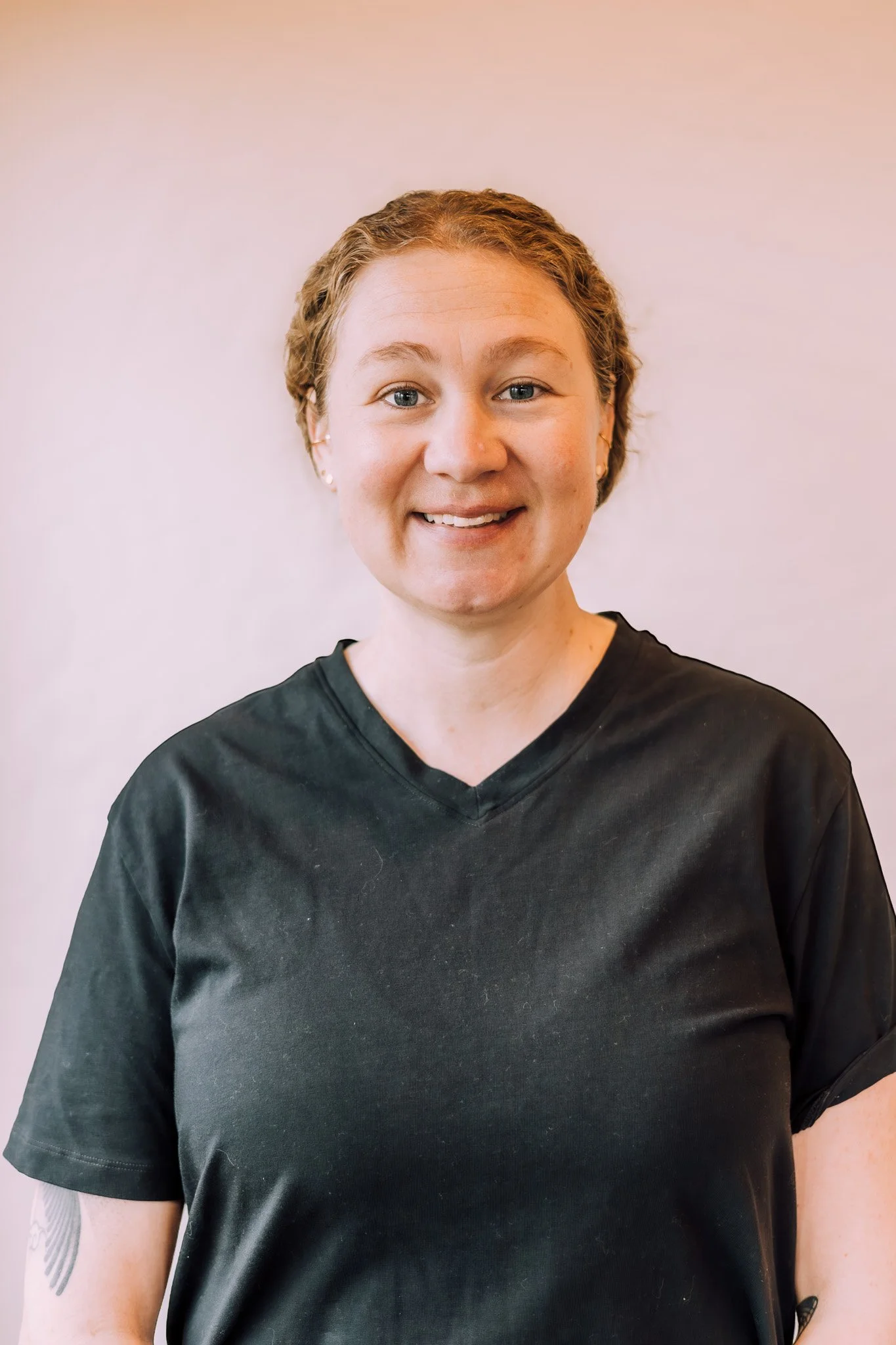 A woman with short, wavy hair smiling, wearing a black V-neck T-shirt, standing against a pale wall.