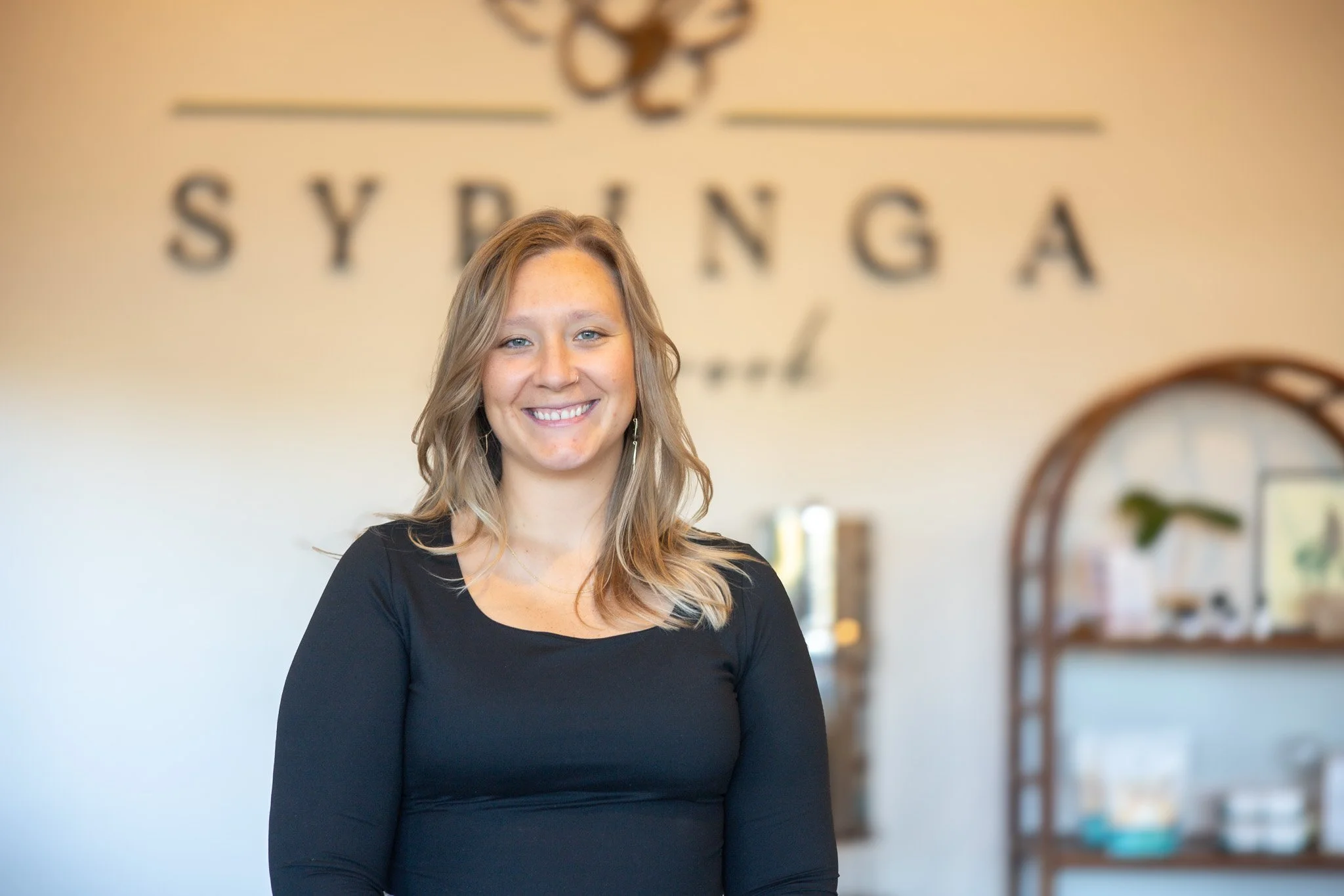 Smiling woman with blonde hair wearing a black top standing in front of a blurred background with the words 'SYRINGA' and a decorative logo on the wall.