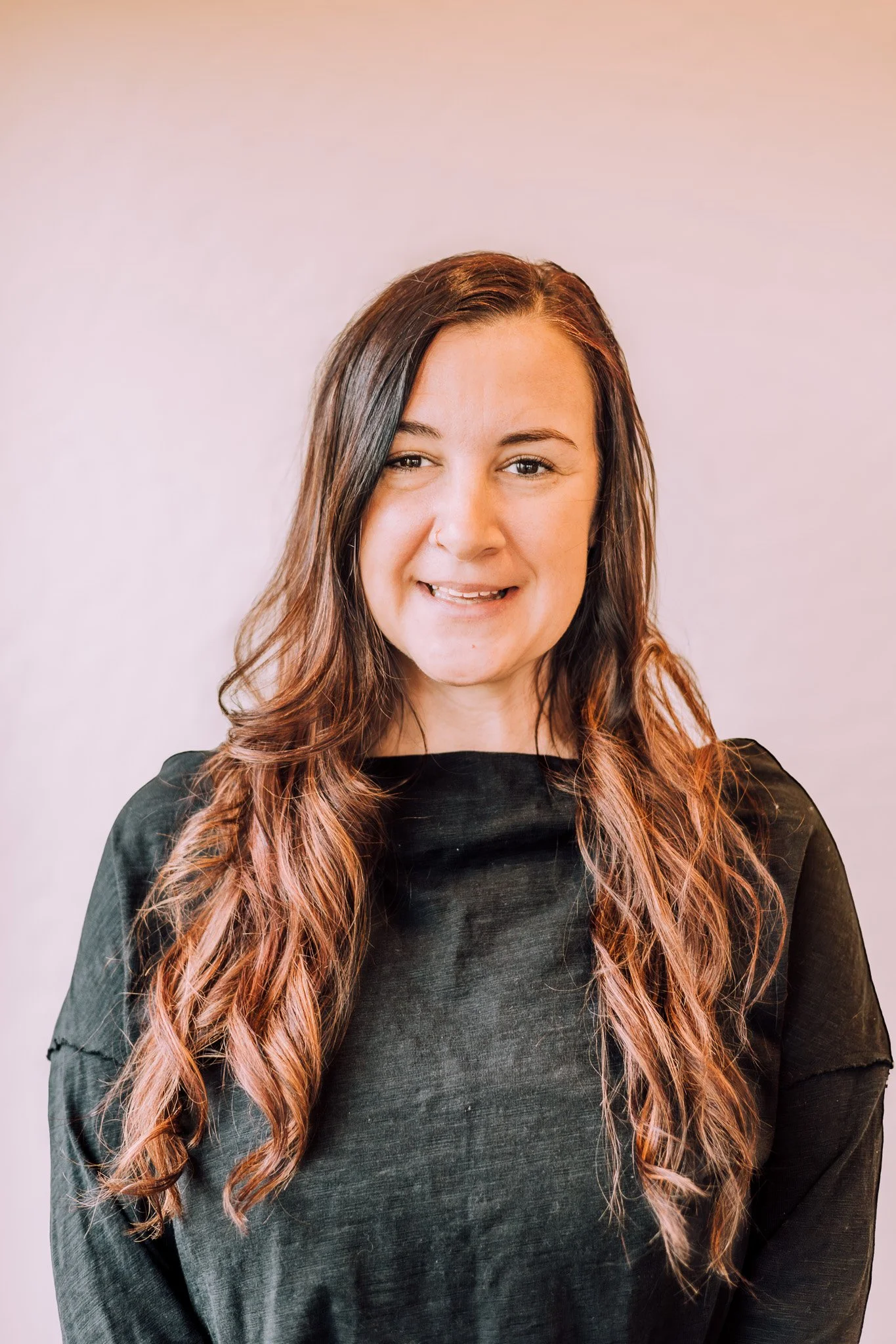 A woman with long wavy hair, smiling, wearing a black top, standing against a light-colored background.