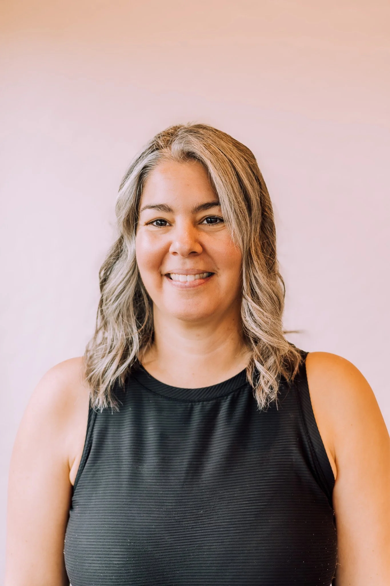 Portrait of a woman with shoulder-length wavy blonde hair, smiling, wearing a black sleeveless top, against a plain light-colored background.