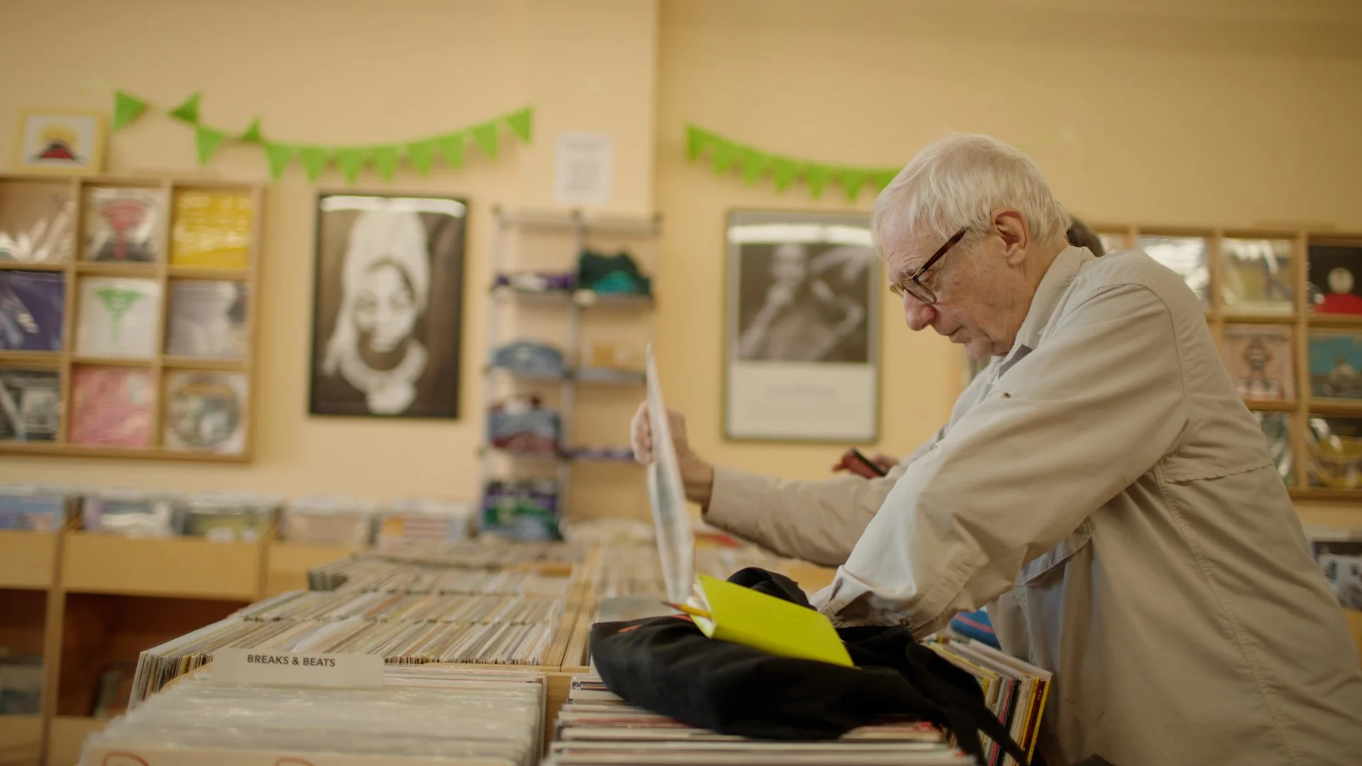 An elderly man with glasses searches through records in a record store, surrounded by shelves of vinyl albums and posters on the walls.