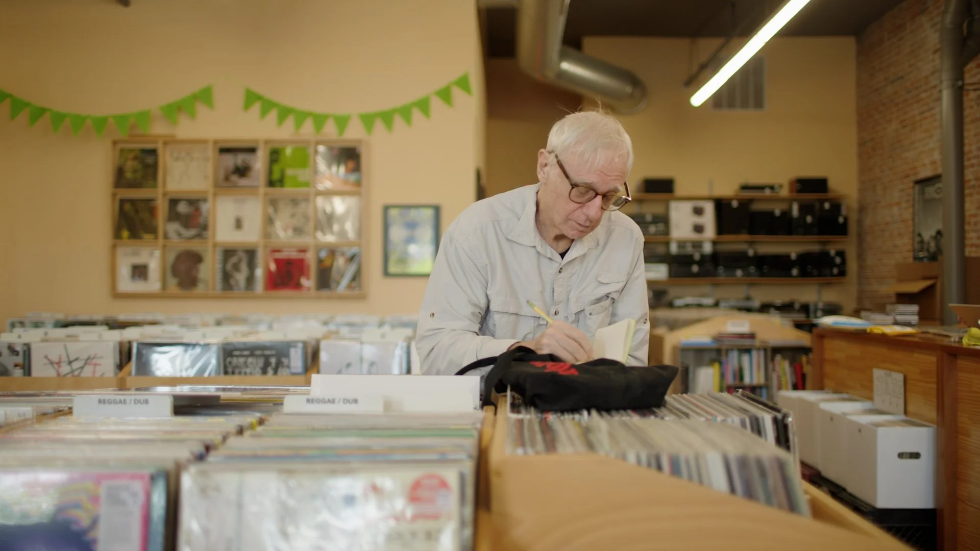 An elderly man with white hair and glasses browsing vinyl records at a record store, taking notes in a notebook.