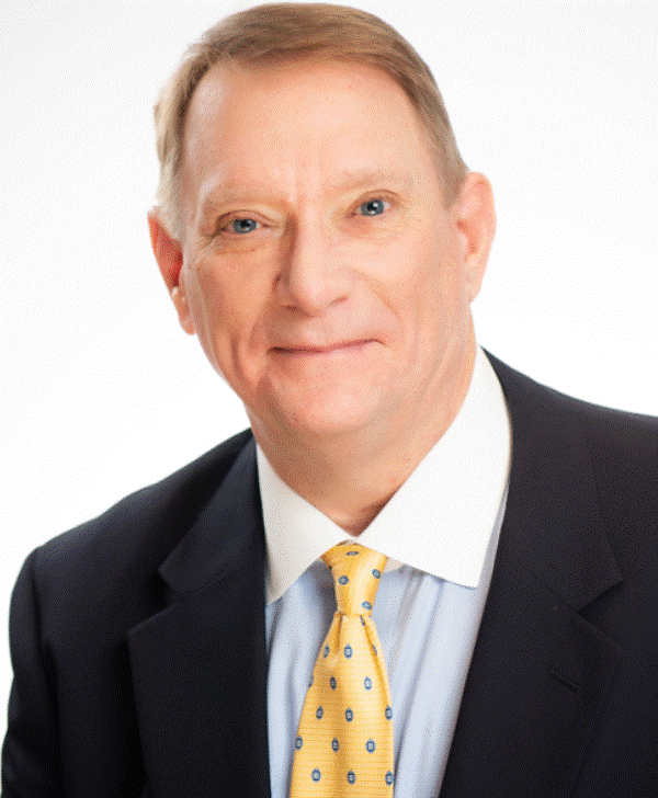 A professional portrait of a middle-aged man with light skin, short light brown hair, wearing a dark suit, white shirt, and a yellow tie with blue dots, smiling slightly against a plain white background.