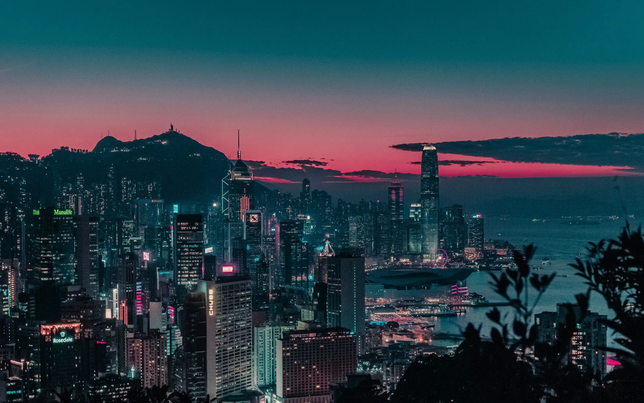 A city skyline during twilight with tall buildings, illuminated signs, and a body of water in the foreground. The sky has hues of pink and blue, with some clouds.