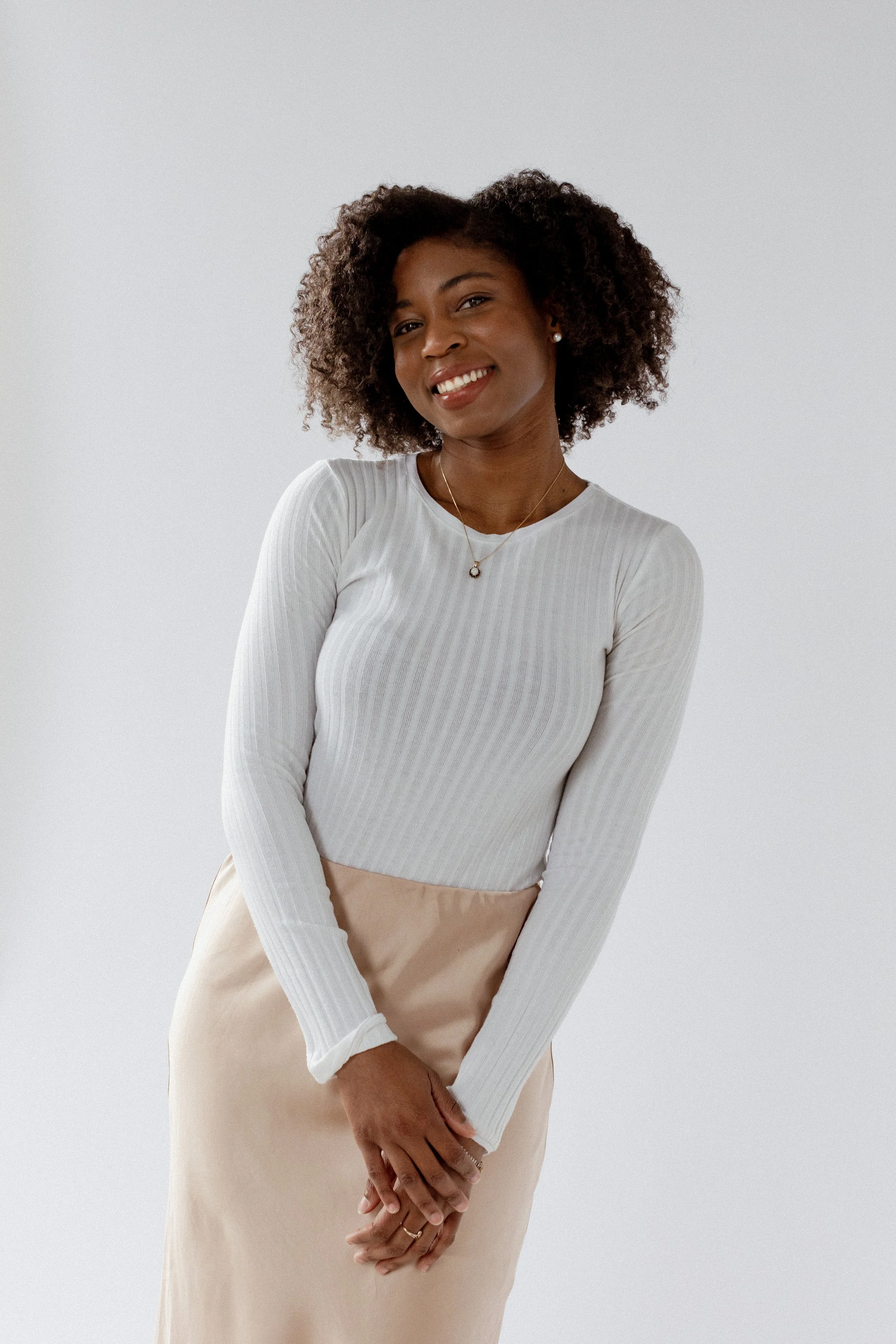 A woman with curly hair wearing a white ribbed long-sleeve top and beige skirt, smiling and standing against a plain light background.