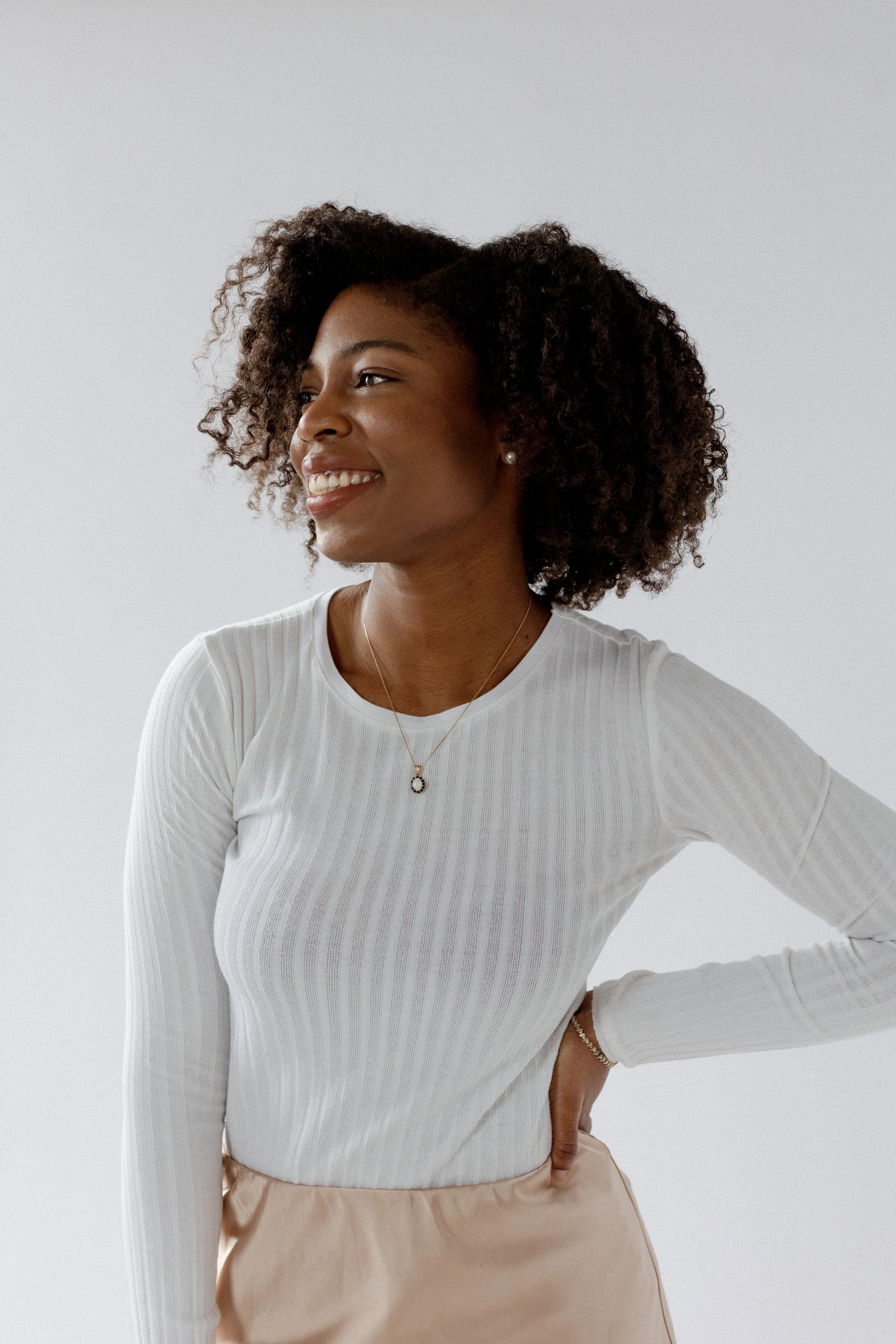 A smiling woman with natural curly hair wearing a white long-sleeve ribbed top and beige skirt, standing against a plain white background.