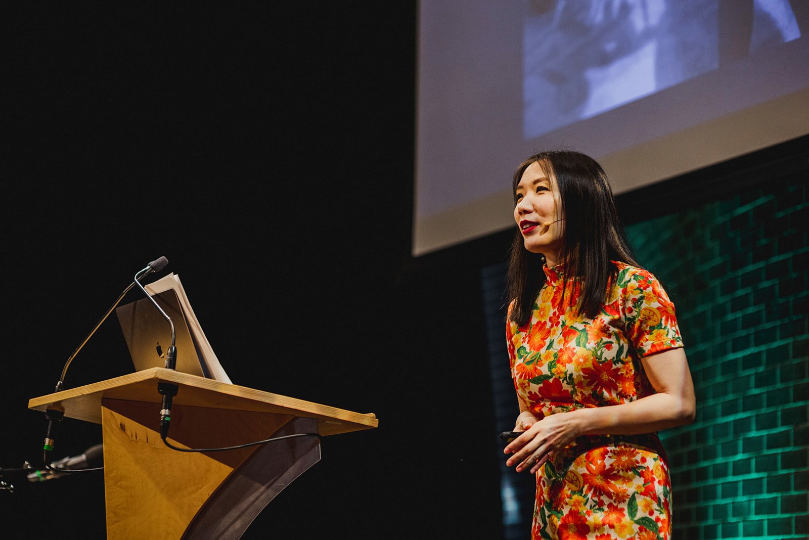 Pennylane Shen at Creative Mornings Vancouver, in the SFU Woodwards Building with a large screen behind her and a wooden podium with a laptop and microphone.
