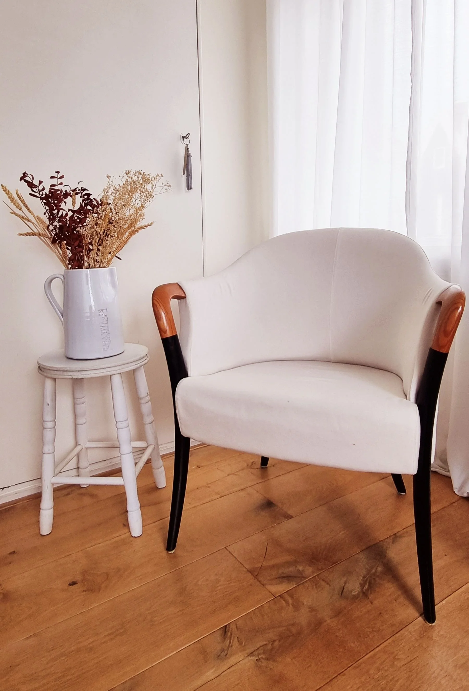 A white armchair with black and wooden legs, placed on a wooden floor. Next to the chair is a small, round, white side table holding a white vase with dried flowers. In the background, there are white curtains and a plain white wall with a small hook and tassel hanging from it.