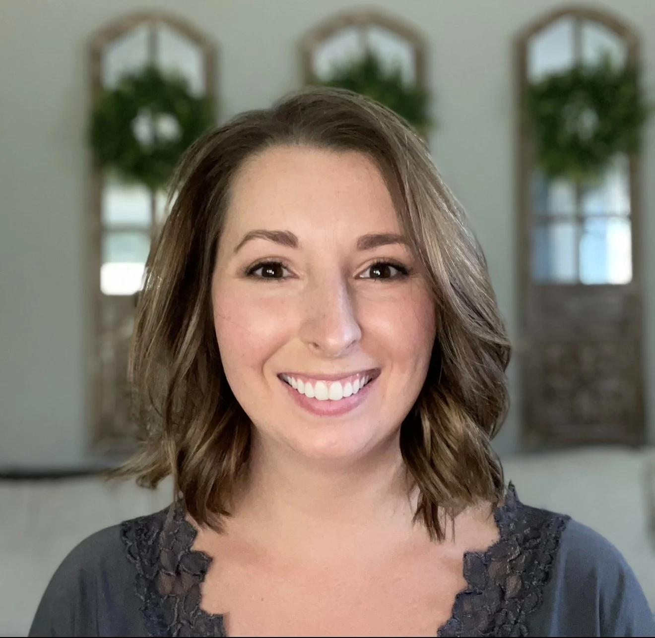 A woman with shoulder-length brown hair smiling at the camera, standing indoors with three decorative windows in the background.