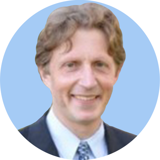 A man with curly light brown hair wearing a suit and tie, smiling against a light blue background.