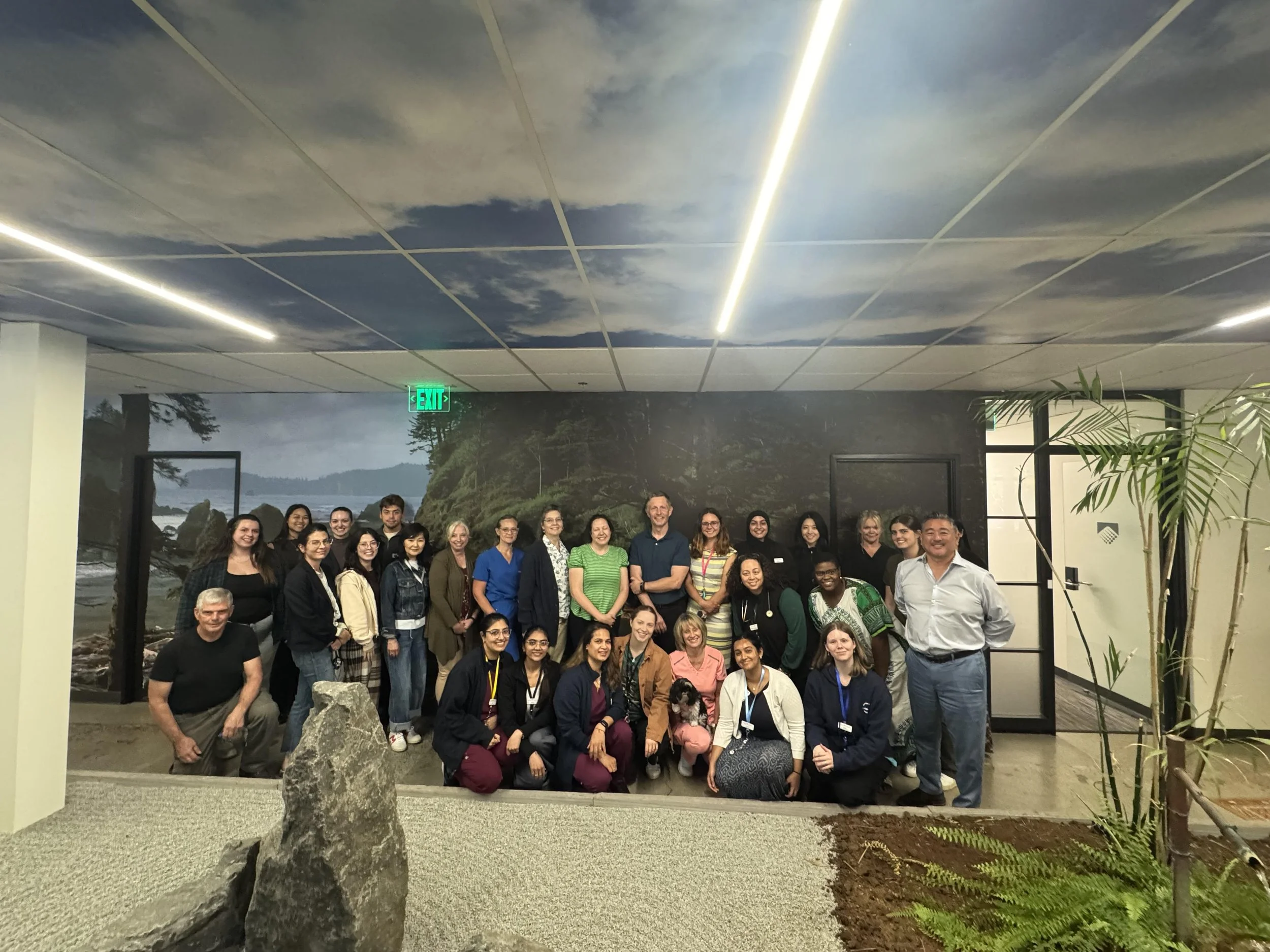 A group of approximately 28 diverse people of various ages and genders posing for a photo indoors, with a mural of a natural landscape on the wall behind them, and plants in the foreground.