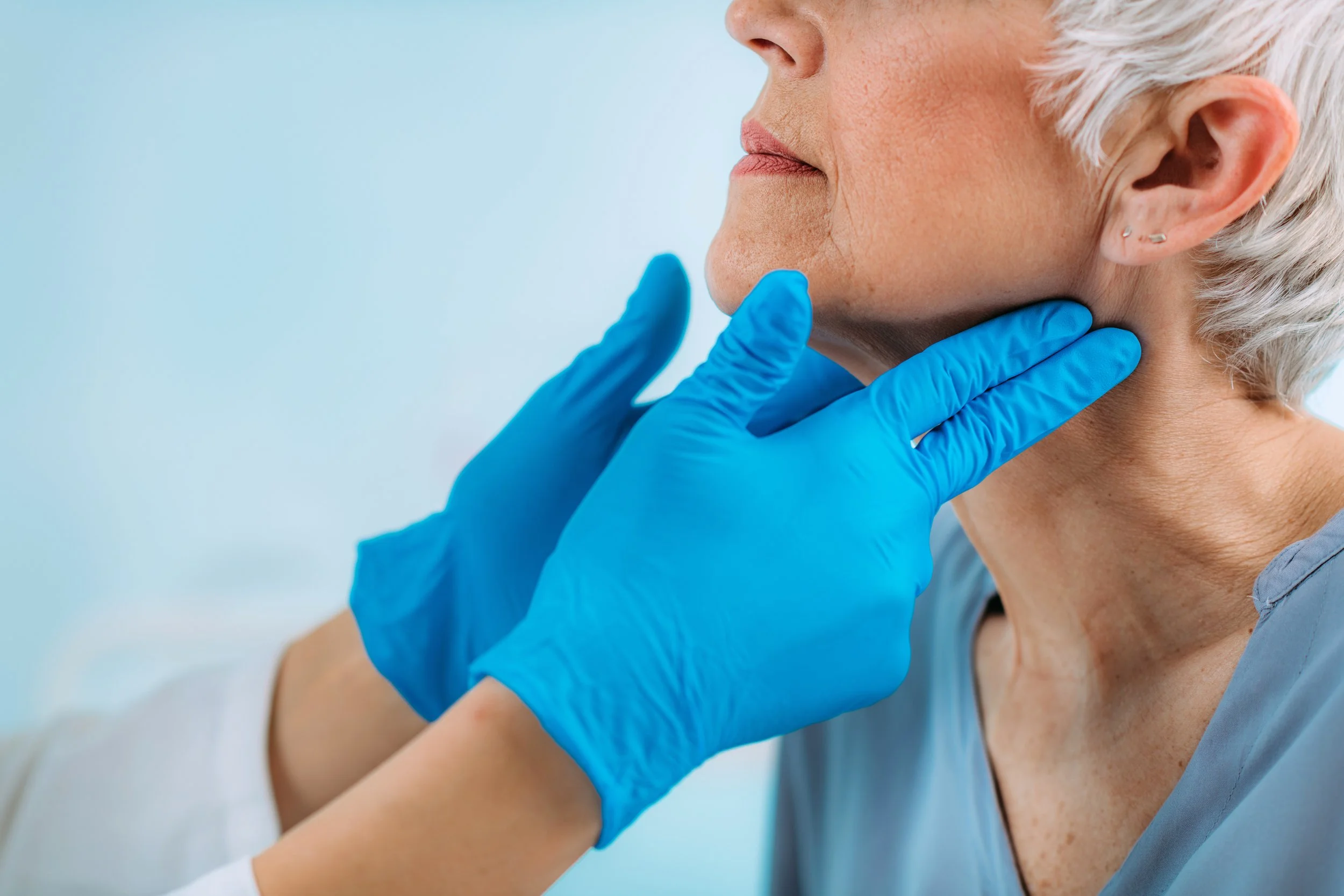 A healthcare professional in blue gloves examining a senior woman's neck in a medical setting.