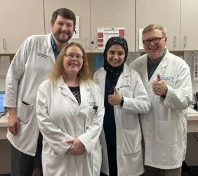 Group of four healthcare professionals, three women and one man, wearing white lab coats, standing in a medical setting, smiling and posing for the photo.