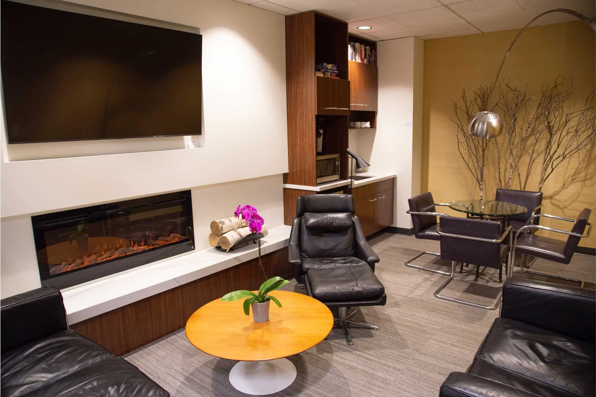 Modern living room with a large flat-screen TV, electric fireplace, black leather chairs, a round wooden coffee table with a potted plant, and a dining area with four black chairs around a glass-top table. Decor includes a branch wall art, a metallic hanging lamp, a wooden cabinet with books, and a small countertop with a microwave and coffee machine.