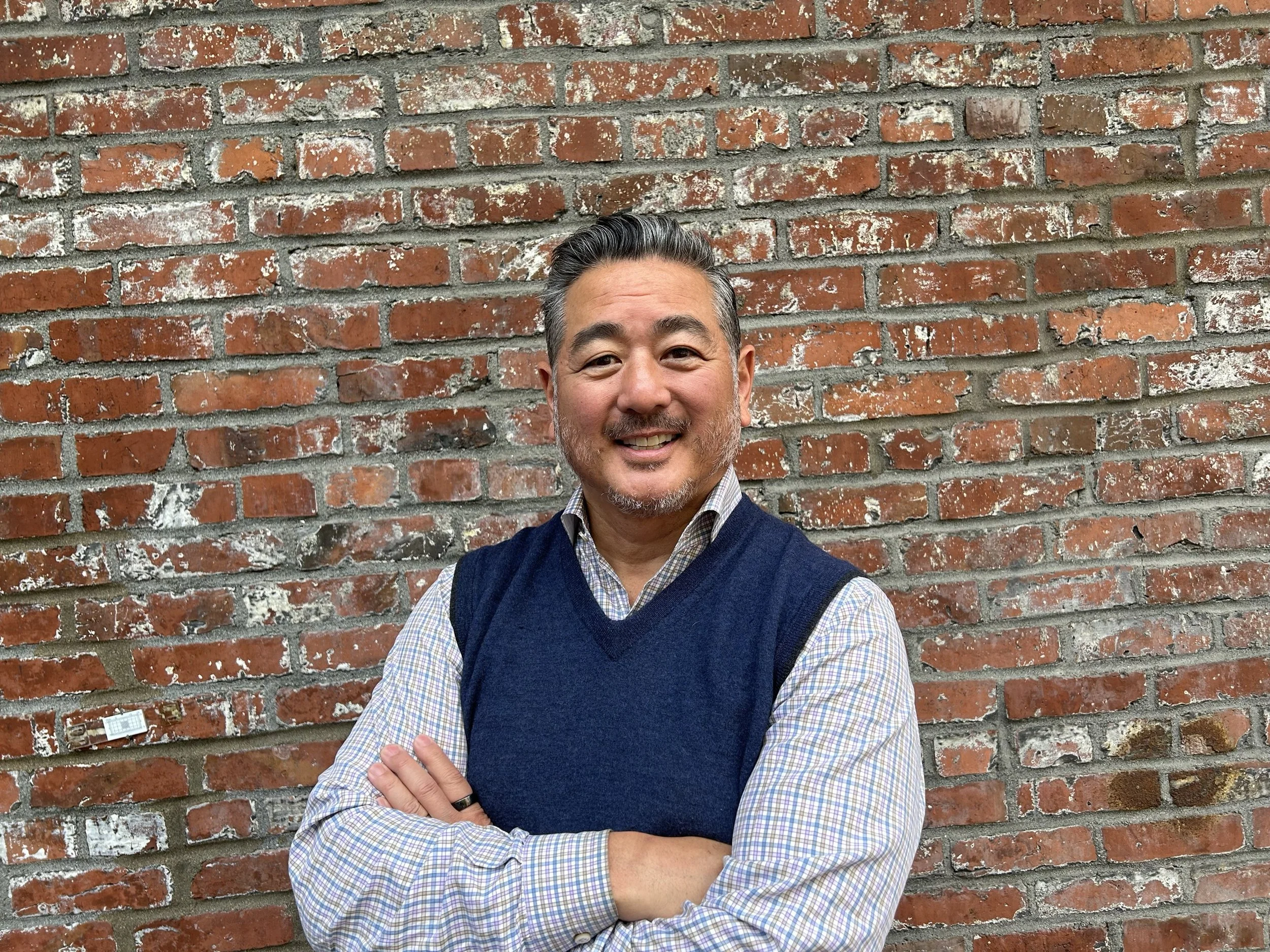 A smiling middle-aged man with gray hair and facial hair, wearing a plaid shirt and navy blue sweater vest, standing with arms crossed in front of a brick wall.