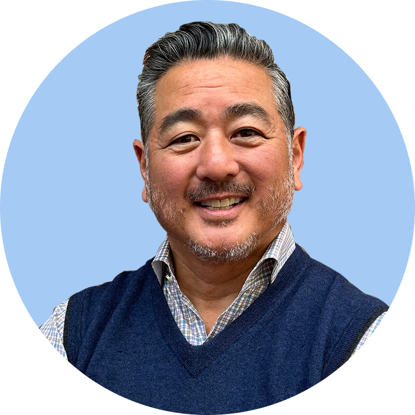 A smiling middle-aged man with gray hair and a beard, wearing a plaid shirt and a navy sweater vest, against a light blue background.