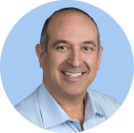 Headshot of a smiling middle-aged man with short dark hair, wearing a light blue collared shirt, against a light blue background.