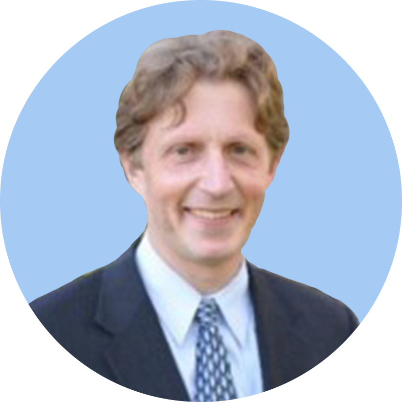 A man with curly brown hair wearing a dark suit, white shirt, and patterned tie, smiling against a blue background.