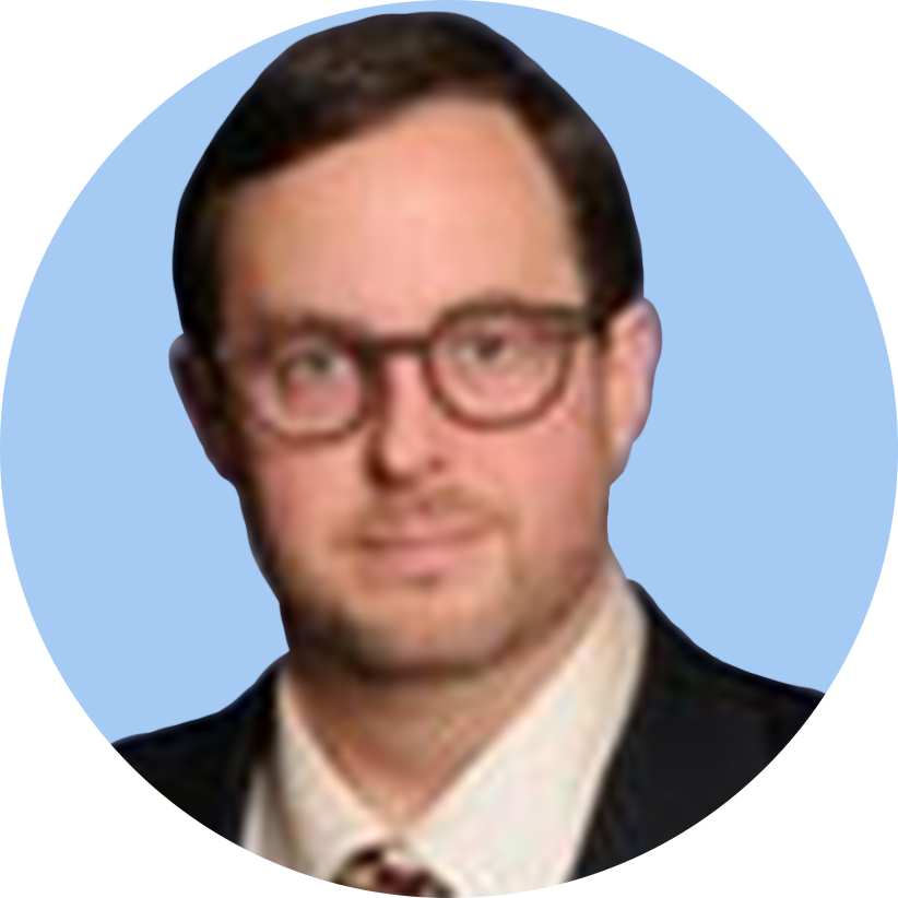 A man with glasses and dark hair wearing a suit and tie, posing against a light blue background.