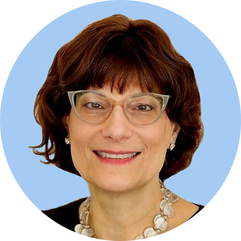 A woman with short brown hair, glasses, wearing pearl earrings and a pearl necklace, smiling against a blue background.