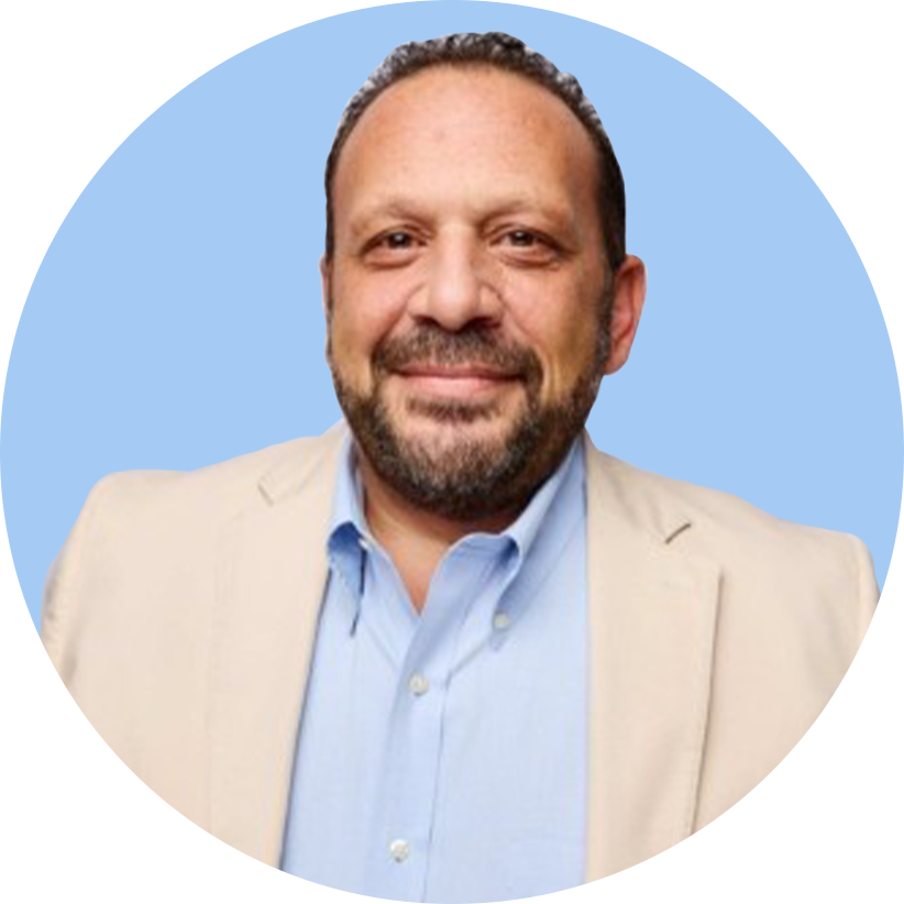 A man with a beard and short hair smiling, dressed in a light-colored blazer and blue dress shirt, against a split light blue and black background.