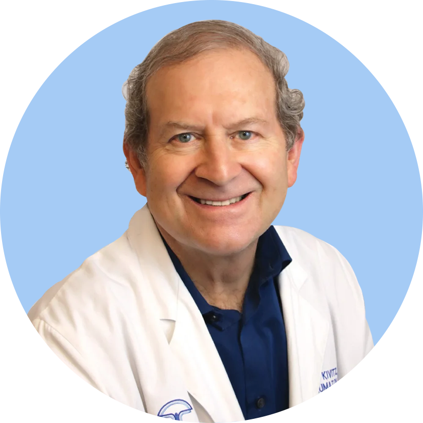 A smiling middle-aged man with gray hair and blue eyes wearing a white lab coat over a dark blue shirt, set against a light blue background with a curved design.