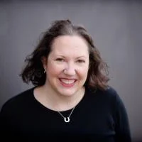 A woman with shoulder-length brown hair and a black top, smiling against a gray background.