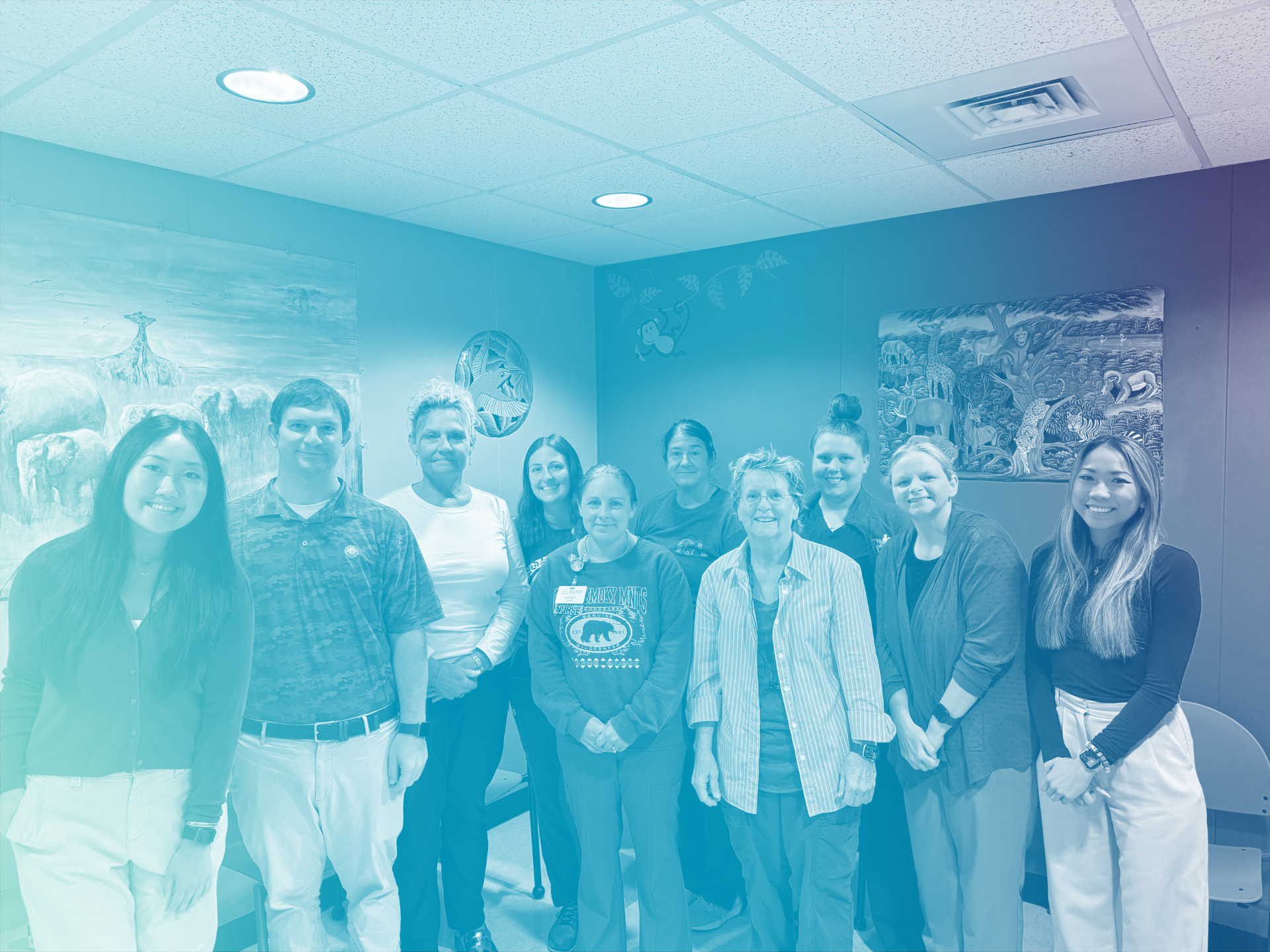 Group of ten women and one man standing together in a room with animal murals on the walls.