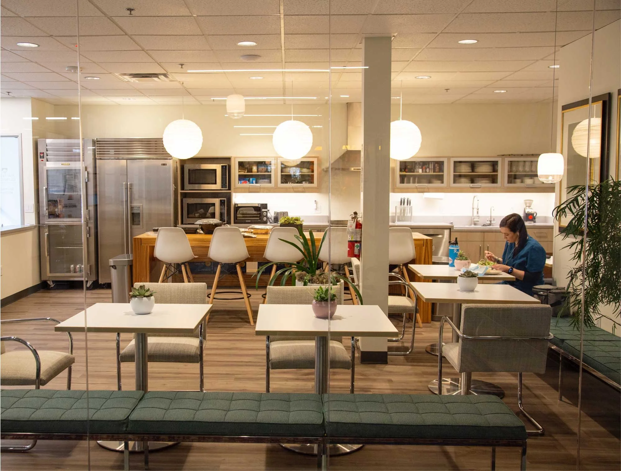 Interior view of a modern cafeteria with a woman eating at a table, white chairs, potted plants, and a fully equipped kitchen in the background, photo taken through glass.