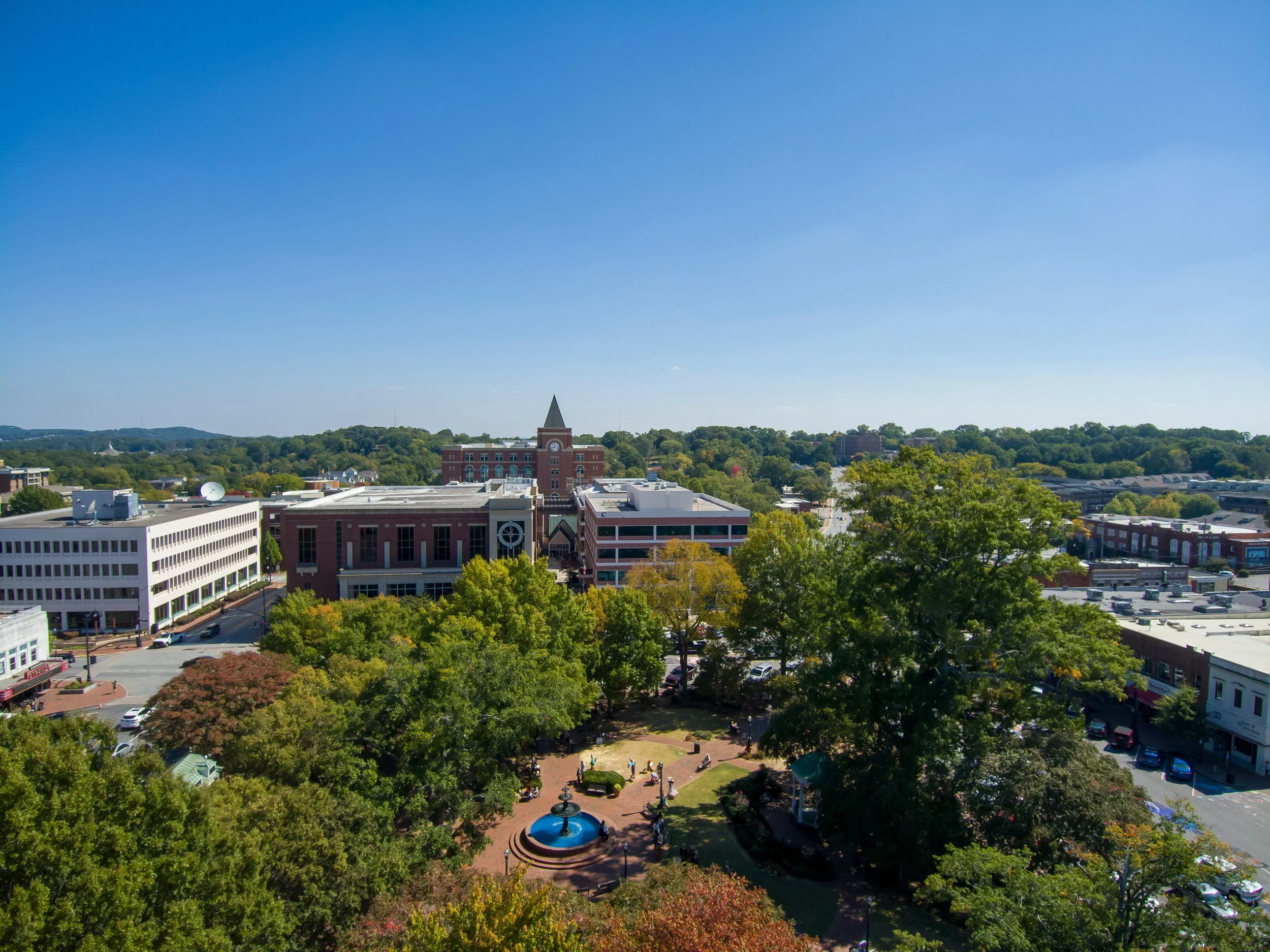 Aerial view of a downtown area with trees, buildings, and a park with a fountain under a clear blue sky.