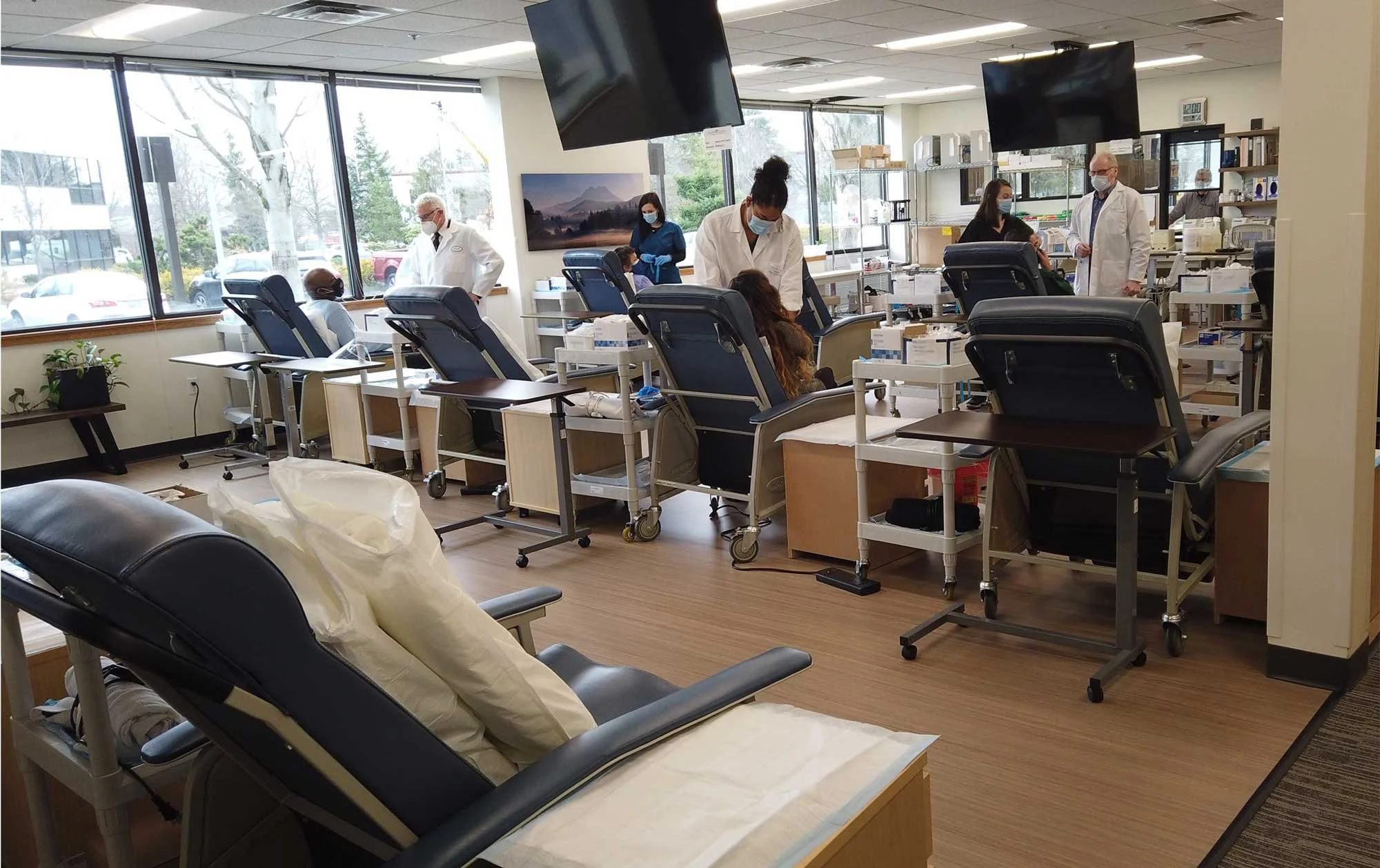 Blood donation center with patients in reclining chairs and healthcare staff assisting, large windows, monitors, and medical supplies.