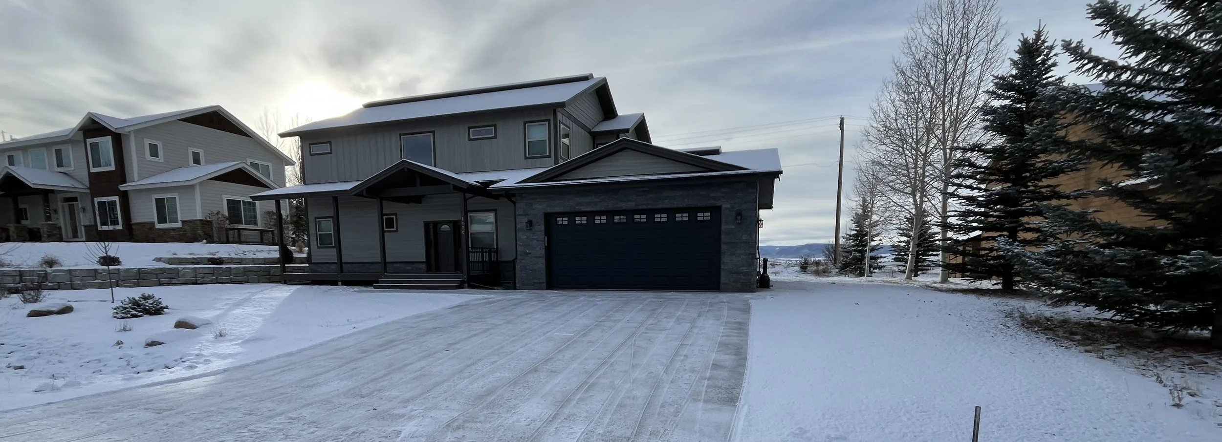 Modern multi-story house with gray and black exterior, stone accents, and a black garage door, situated in a snowy landscape with trees and neighboring houses visible in the background.