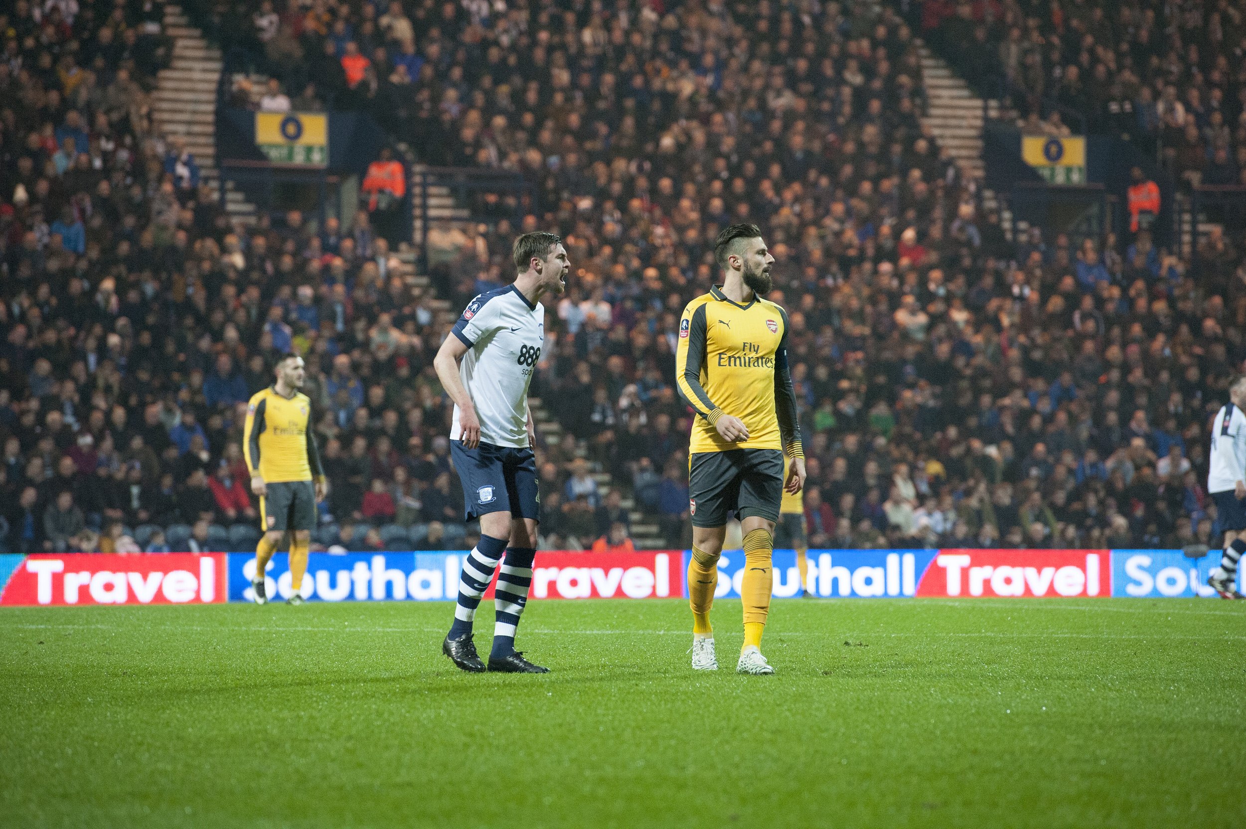 Soccer players on the field during a match, with one wearing a white and navy uniform and the other in a yellow and black kit, in front of a crowd at stadium.