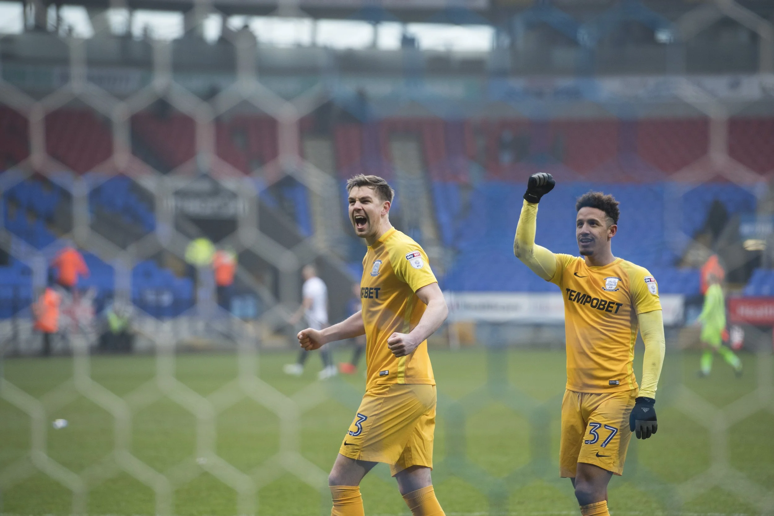 Two soccer players in yellow jerseys celebrating on the field, seen through a goal net. The player on the left is clenching his fists with a joyful expression; the player on the right is raising his fist in celebration. The background shows a stadium