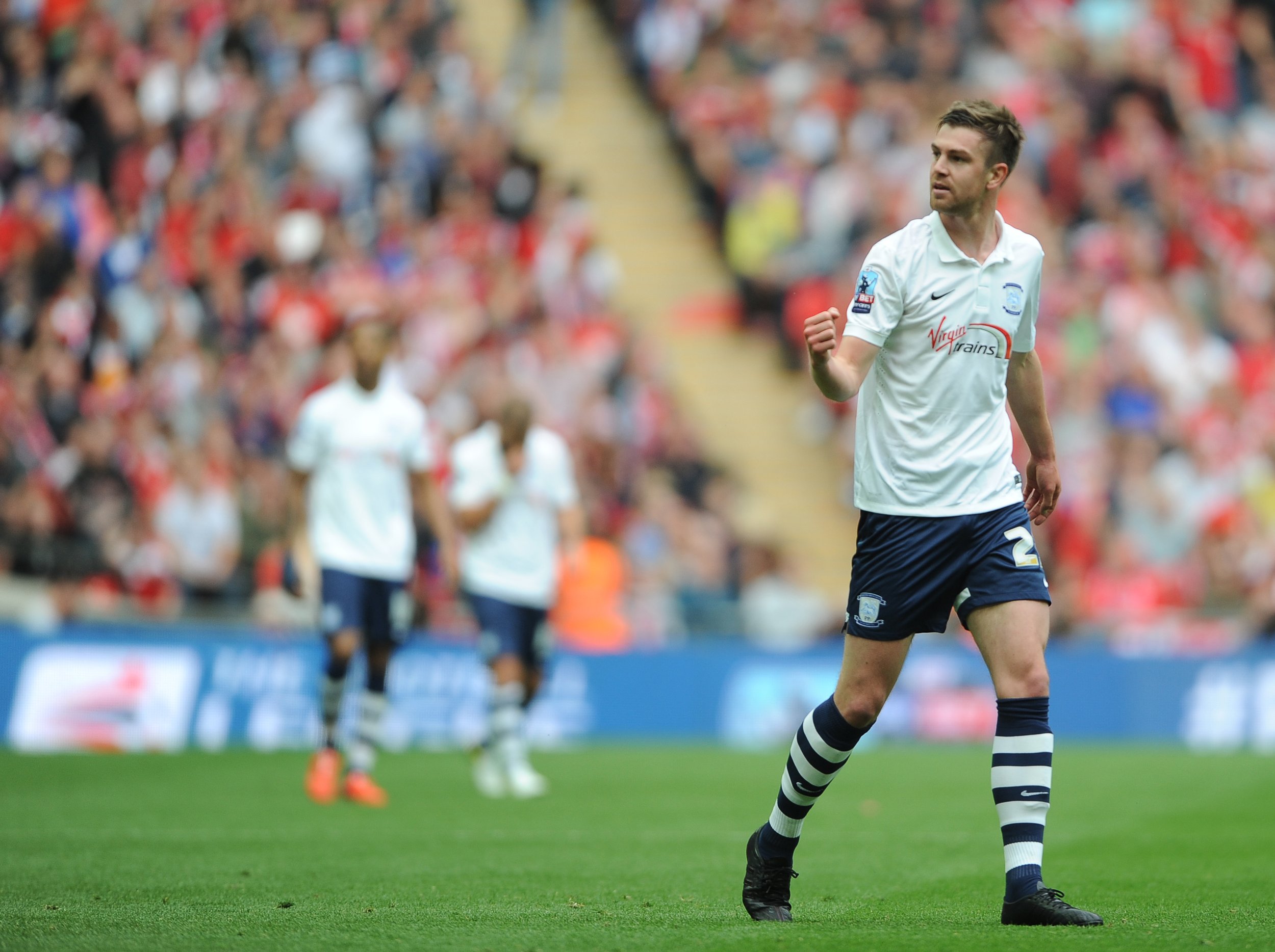 A soccer player in a white jersey celebrating on the field with clenched fist, with a crowd in the background.
