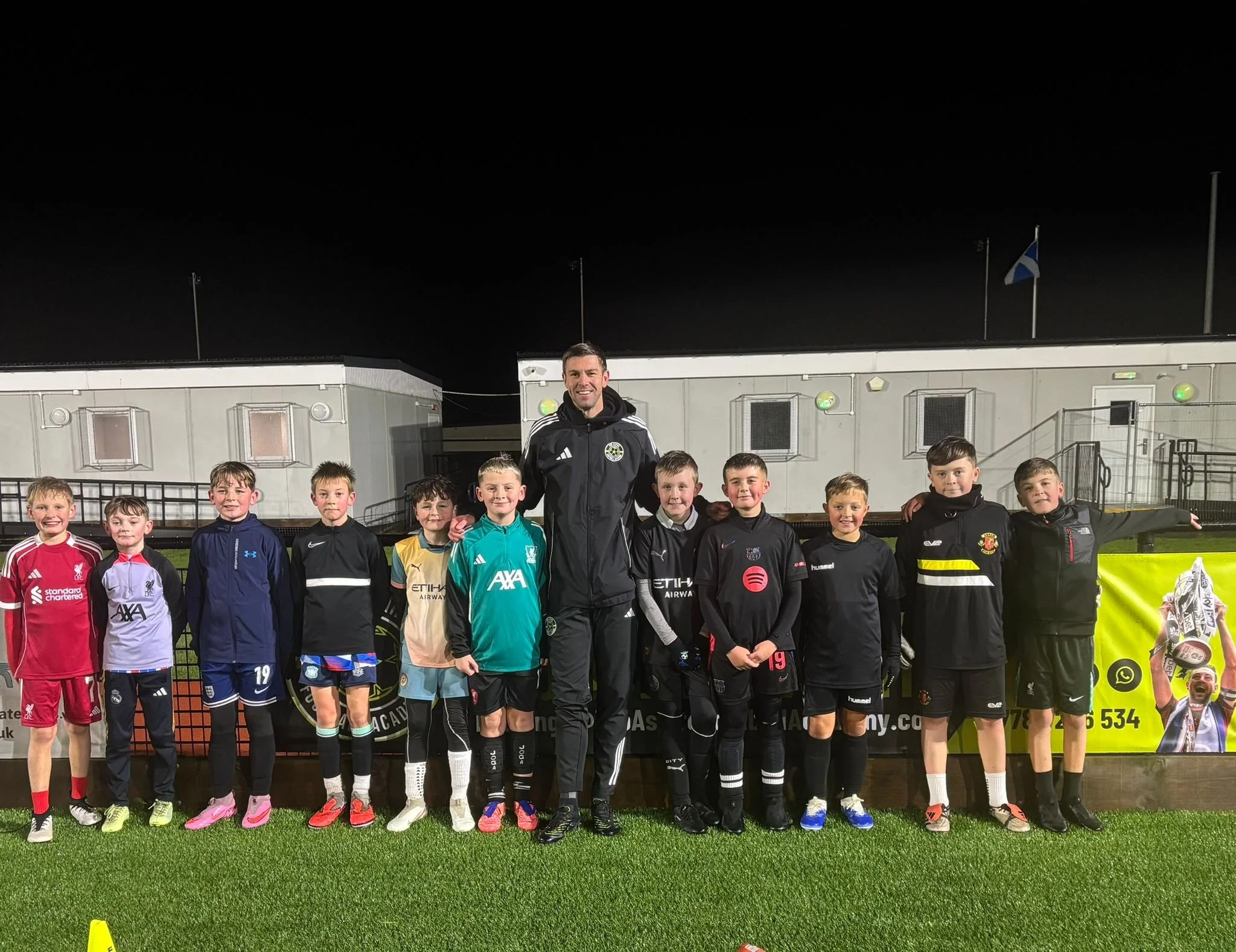 A coach and two young boys on a soccer field, practicing with a soccer ball. The coach is guiding the boy who is kicking the ball, while the other boy stands near the goal. There are yellow training cones on the field, and a fenced area surrounds the field with houses in the background.