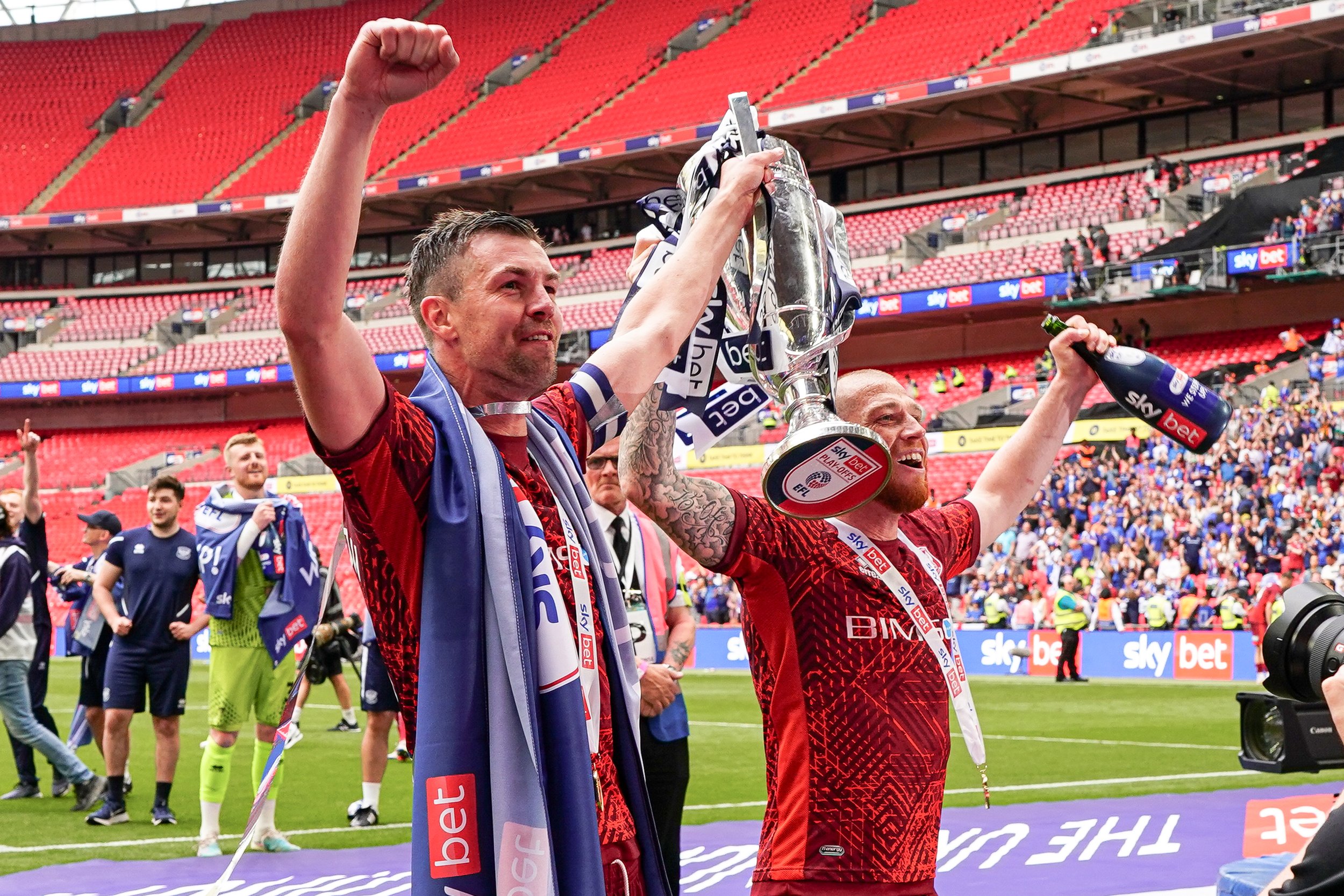 Two soccer players celebrating on the field, holding a trophy, with a stadium filled with spectators in the background.