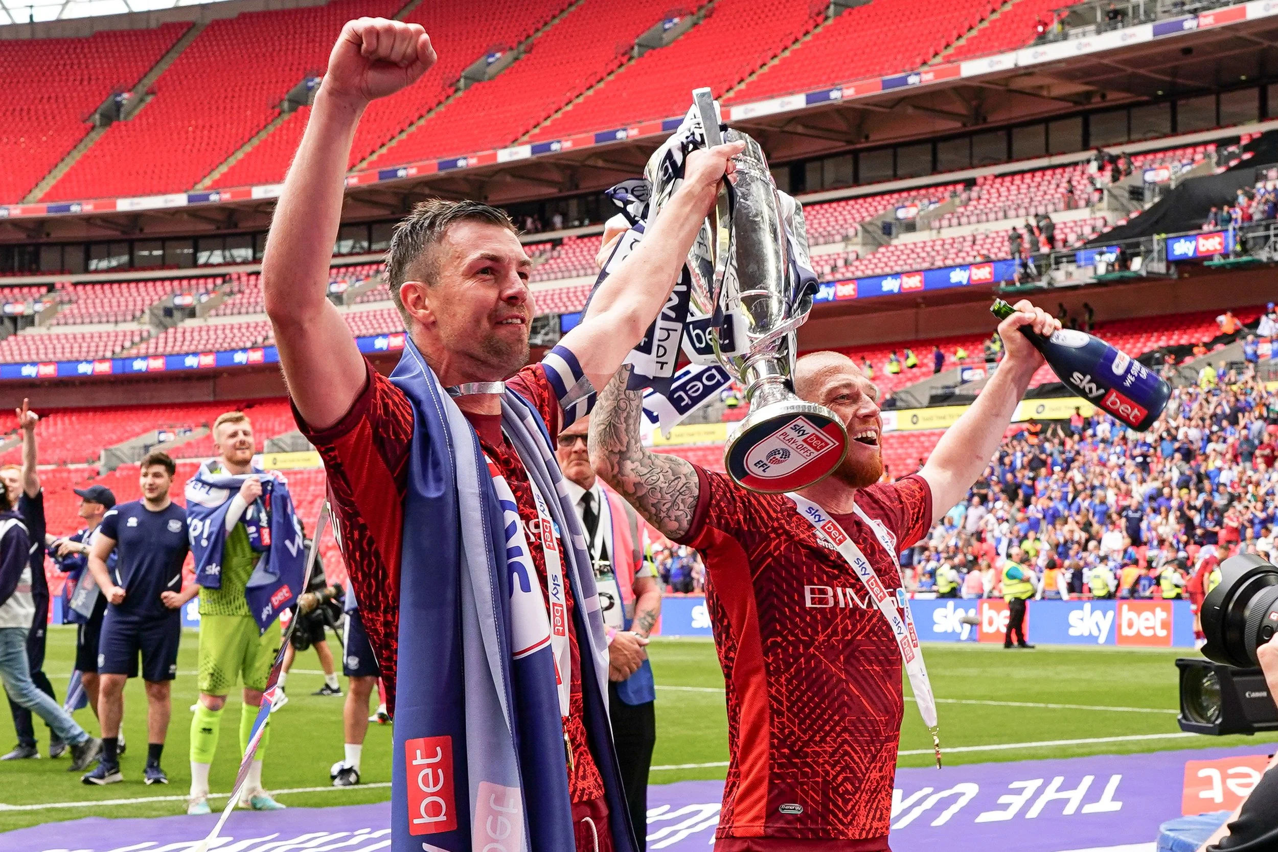 Two soccer players in red jerseys celebrating on the field with a trophy and drinks, surrounded by fans and staff after a match at a stadium.
